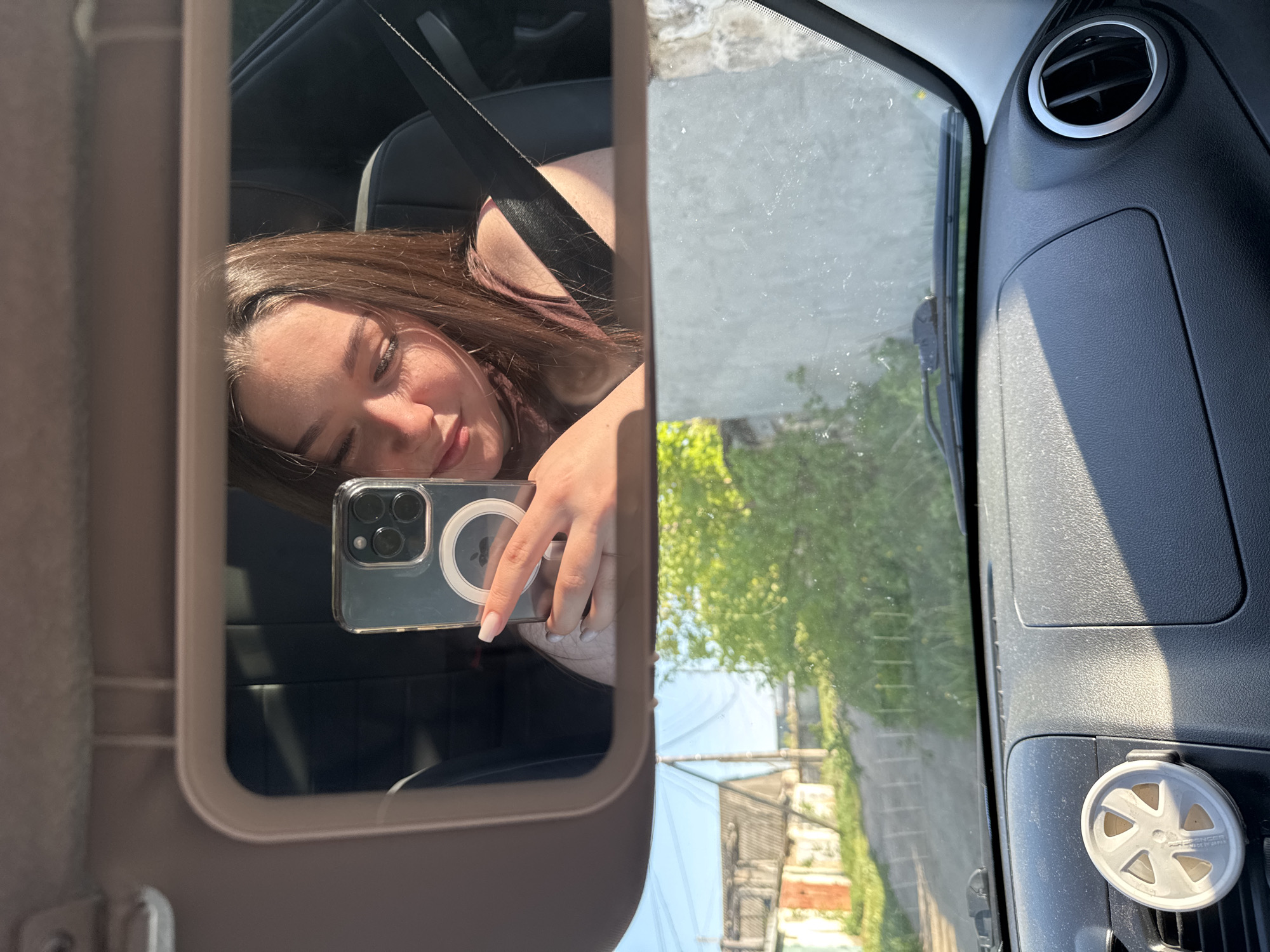 A woman is taking a selfie in the vanity mirror of a car. The mirror reflects the woman's face, wearing a seatbelt. Her left hand holds a smartphone. Outside the car window, the reflection shows a streetscape with trees and buildings. The location is Гамово, Russia. The car's dashboard and air vent are visible.