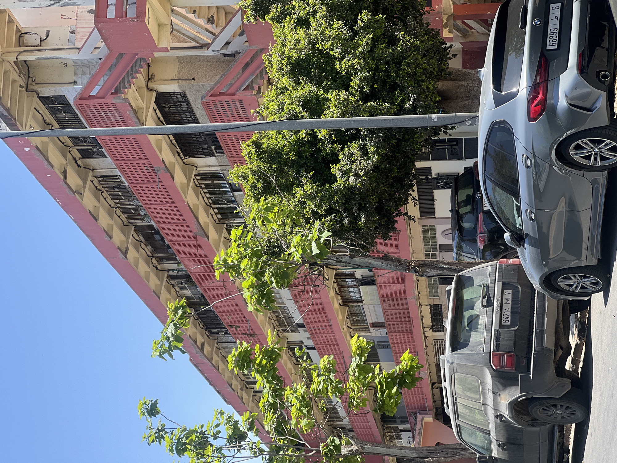 This image captures a vibrant urban scene in Casablanca, Morocco, featuring a multi-story residential building and parked cars under a clear blue sky.
The central subject is an apartment building characterized by its distinct salmon-pink balconies and cream-colored facade. The building rises at least five visible stories, with each floor featuring a continuous balcony bordered by perforated, vertically patterned salmon-pink railings. Windows are fitted with black security bars, and external utility conduits and boxes are visible on the facade. On the far right, an external concrete staircase, matching the salmon-pink color scheme, ascends the building. The architecture appears somewhat weathered, consistent with an older urban structure.
In the foreground, two primary vehicles are parked. On the left, a dark gray Toyota Land Cruiser Prado, showing signs of dust accumulation, is parked facing away from the viewer. Its Moroccan license plate reads "64214 | 7". To the right of the Toyota, a cleaner, gray BMW 1 Series hatchback is parked, also facing away, displaying the Moroccan license plate "66899 ط 6". A portion of another dark SUV is partially visible behind the Toyota.
Numerous trees with dense green foliage are dispersed throughout the scene, partially obscuring parts of the building and the vehicles. A prominent, tall, gray utility pole stands vertically in the mid-ground, positioned slightly right of the center. The sky is bright and cloudless, indicating a sunny day. No individuals are visible within the frame. The cars are parked on a paved street or alongside a curb.
