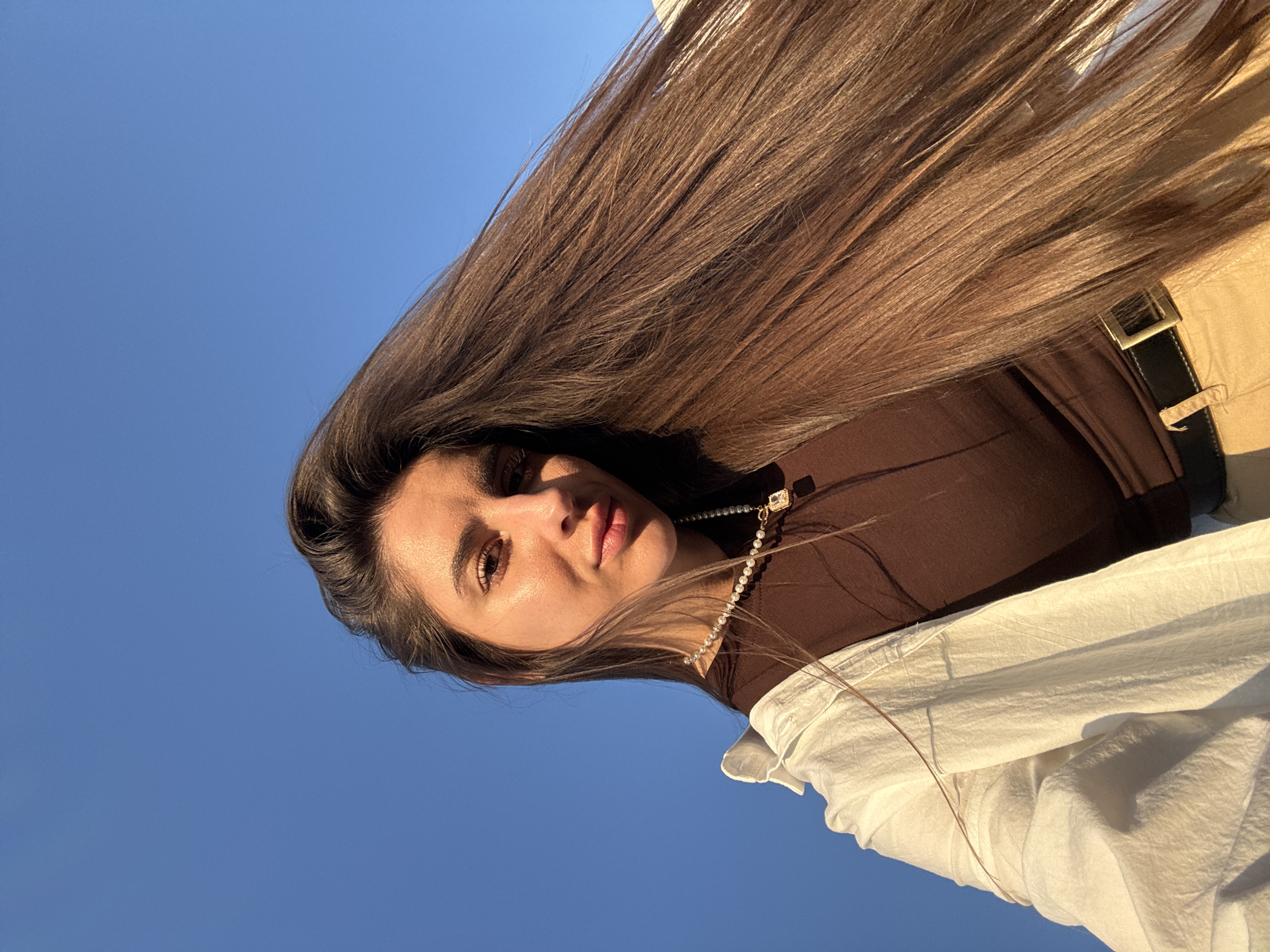 The media file displays a close-up, low-angle shot of a young woman against a clear blue sky. She is positioned in the lower right quadrant of the frame, with her head tilted upwards. Her long, dark brown hair is fanned out dramatically across the upper portion of the image, catching the sunlight. She is wearing a brown top with a pearl necklace and a white open shirt draped over her shoulders. Her facial features are visible, with her eyes open and looking towards the camera, her lips slightly parted.
The scene appears to be outdoors, given the bright sunlight and the expansive blue sky. The lighting suggests it is either early morning or late afternoon, with the sun casting a warm glow on her skin and hair. The overall mood is bright and perhaps contemplative or playful, with the dynamic spread of her hair creating a sense of movement. No text is visible in the image. The context provided suggests this image was taken in Tolyatti, Russia.