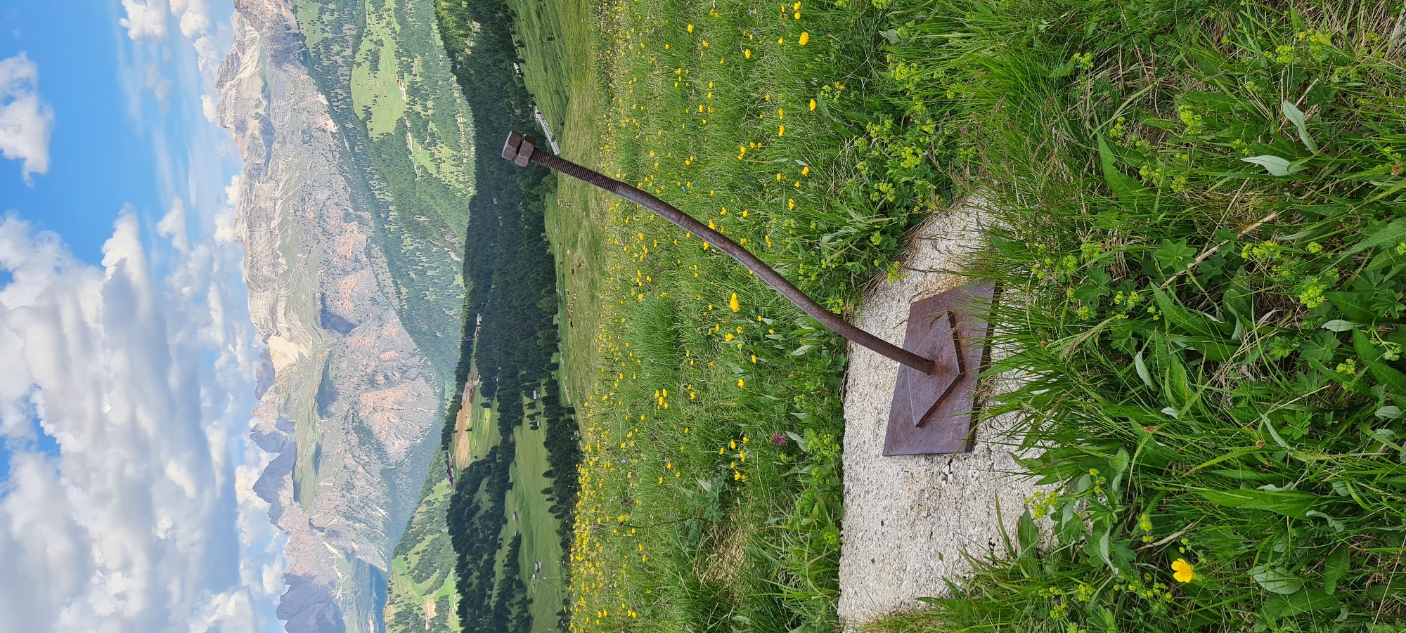 An abstract sculpture, resembling a bolt with two nuts, stands in a grassy, mountainous landscape. The sculpture is made of rusted metal, with a square base and a long, curved rod topped with two hexagonal nuts. It is positioned on a concrete pad surrounded by lush green grass dotted with small yellow flowers. In the background, a vast expanse of rolling hills and rugged mountains unfolds under a partly cloudy blue sky. The mountains exhibit a rugged, rocky texture with patches of snow near their peaks, and are covered in dense evergreen forests on their lower slopes. Further in the distance, there are signs of human activity, including buildings and ski lifts, suggesting a resort area. The overall scene depicts a blend of natural beauty and human intervention in a high-altitude environment, likely in the Dolomites region of Italy, near Sëlva - Wolkenstein - Selva di Val Gardena. There are no people visible in the image.
