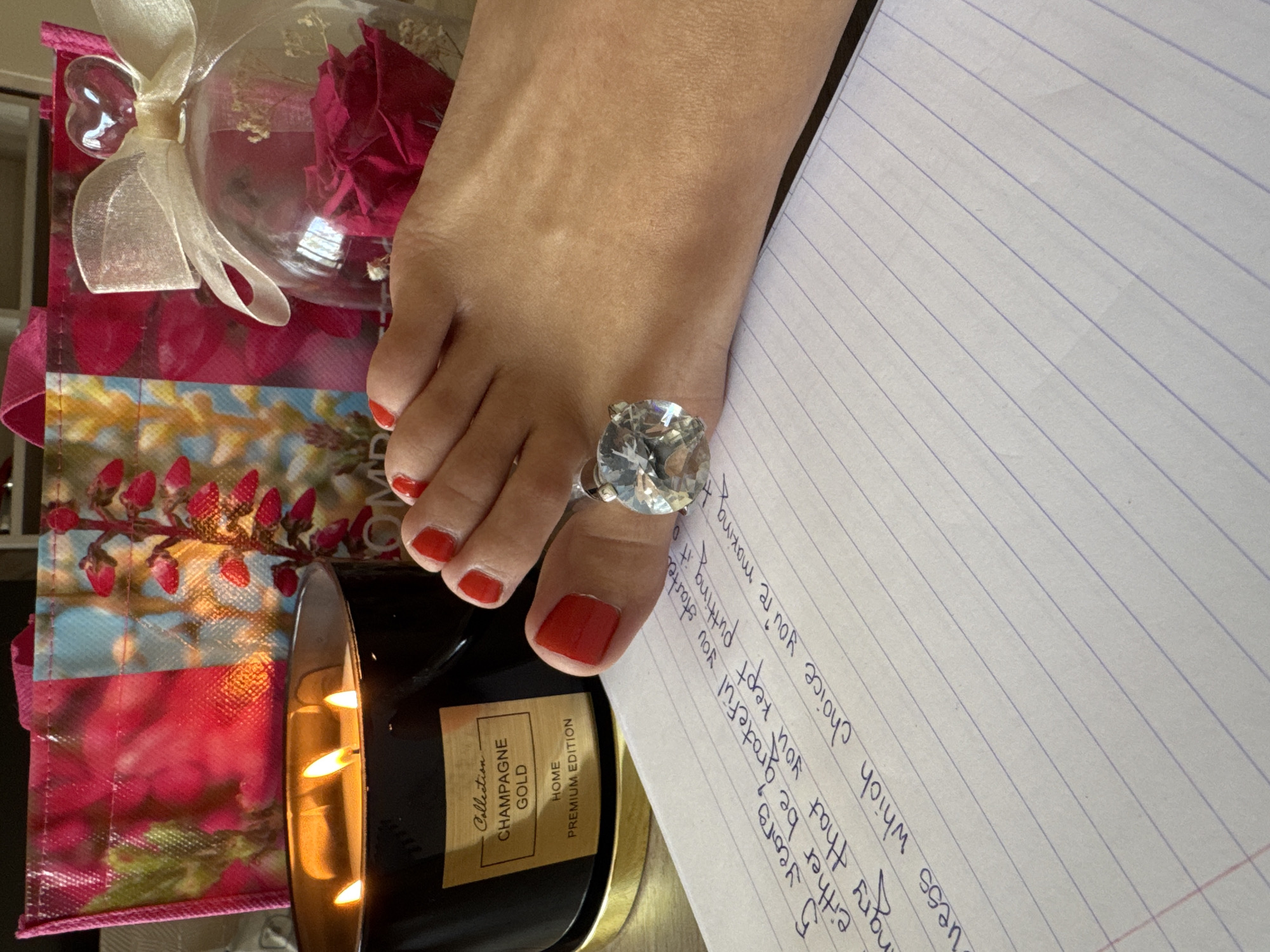 Here is a forensic description of the image:
A photograph features a human foot with red nail polish on the toes, adorned with a large, clear gemstone ring on the second toe. The foot rests on a sheet of lined paper with handwritten text. Beside the foot is a lit candle in a black jar labeled "Collection CHAMPAGNE GOLD HOME PREMIUM EDITION," and a pink floral gift bag with a decorative glass sphere and ribbon. The location appears to be indoors, likely on a desk or table. The specific city and region are indeterminable from the given image.