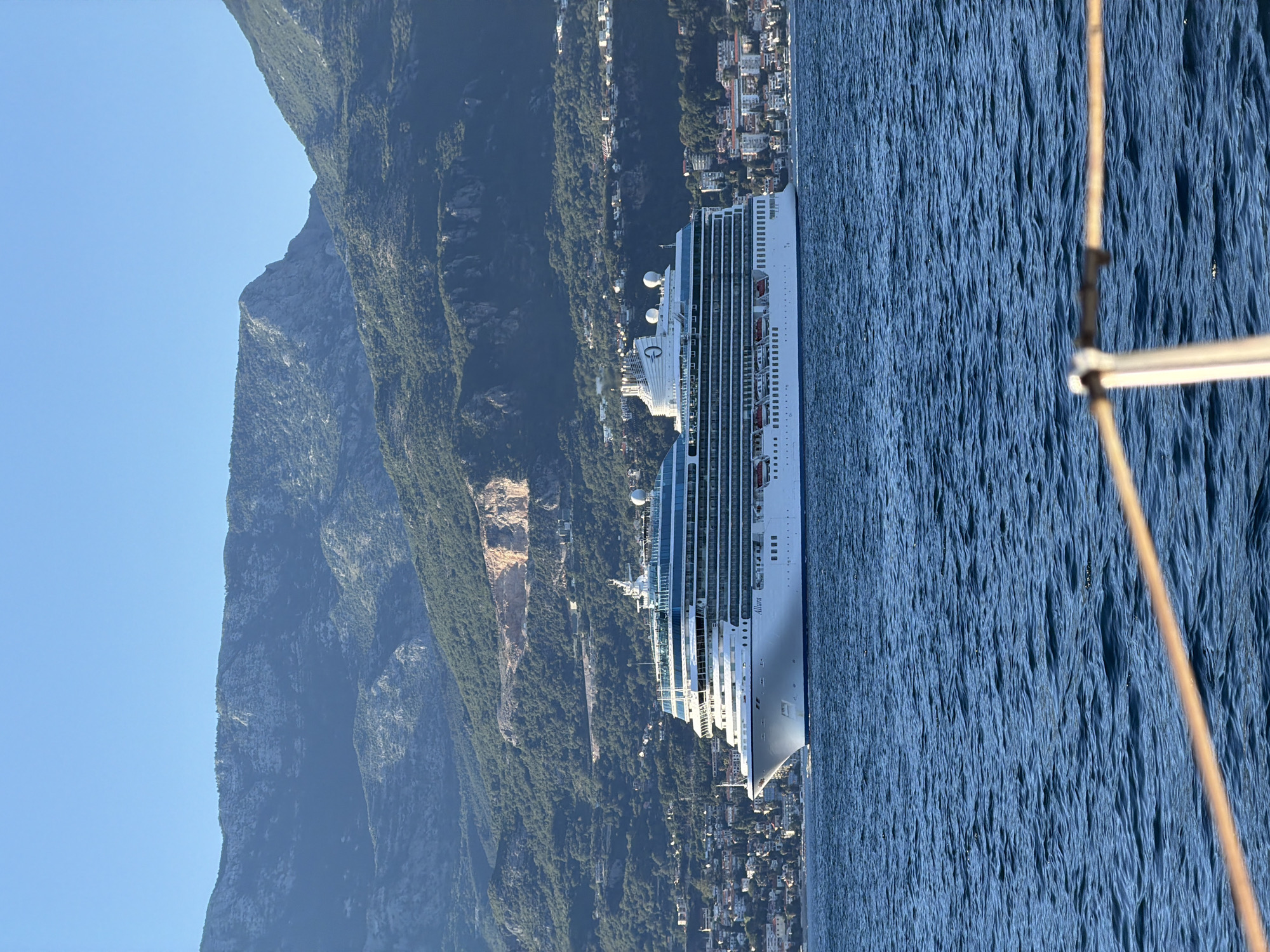 An overcast, clear day captures a large, white cruise ship named "Allura" sailing on calm blue water. The ship is positioned in the middle ground of the image, with its bow facing left. The ship is multi-decked with many windows, and its upper decks are a light blue color. In the background, a densely populated coastal city with buildings of various sizes and colors is visible, nestled at the base of a rugged, green mountain range. The mountains are steep and rocky, with some bare patches of earth, suggesting quarrying or landslides. The sky is a clear, pale blue. In the foreground, a portion of a boat, with ropes and railing, is visible, indicating the photograph was taken from another vessel.