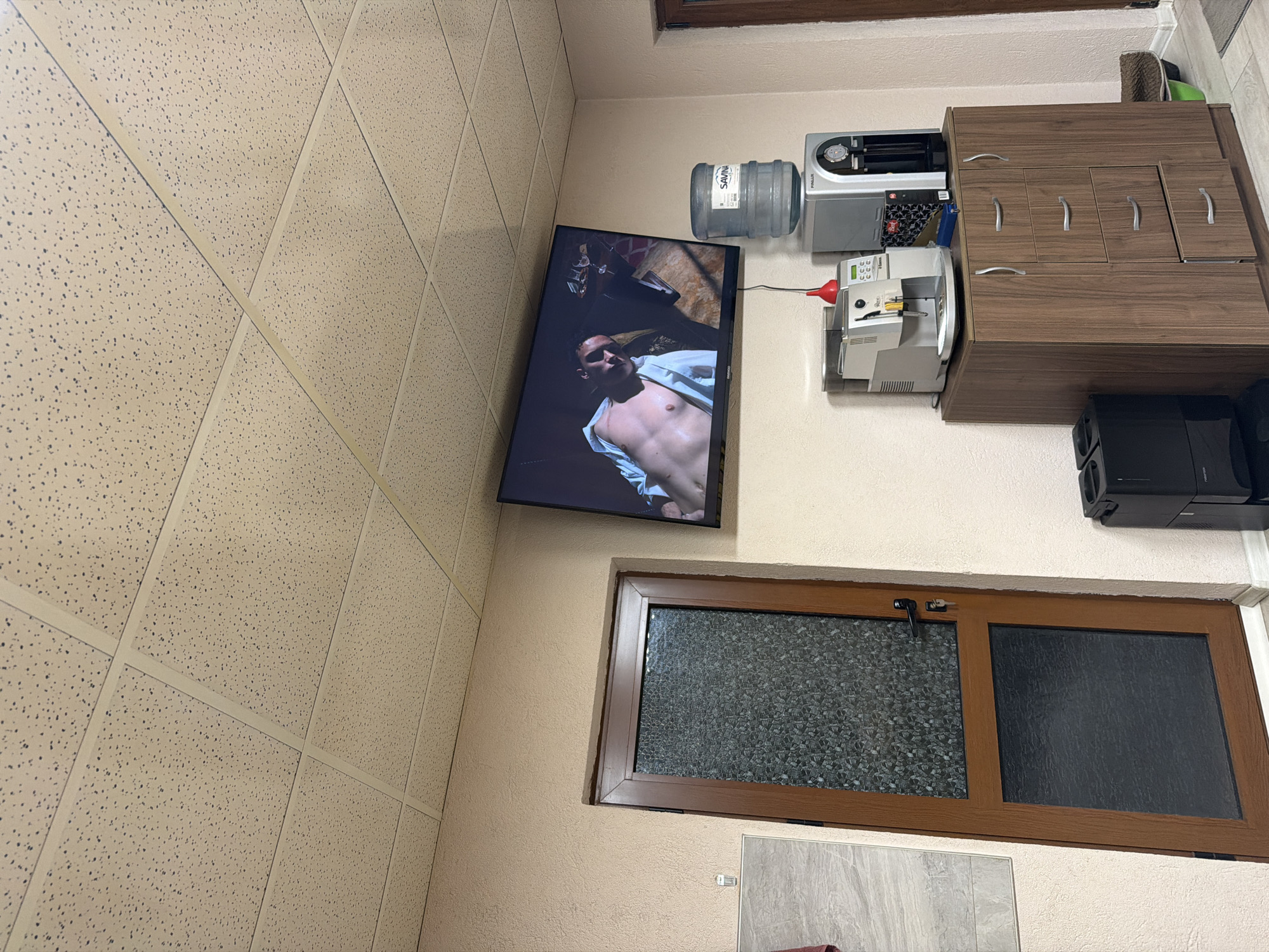 The image shows a room with a television mounted on the wall, displaying a shirtless man. Next to the television is a water dispenser and a coffee machine, placed on a wooden cabinet. Below the television is a window with frosted glass on one side. The wall is painted in a light beige color, and the ceiling is covered with acoustic tiles. A speaker system is positioned adjacent to the wooden cabinet. The location is unspecified.