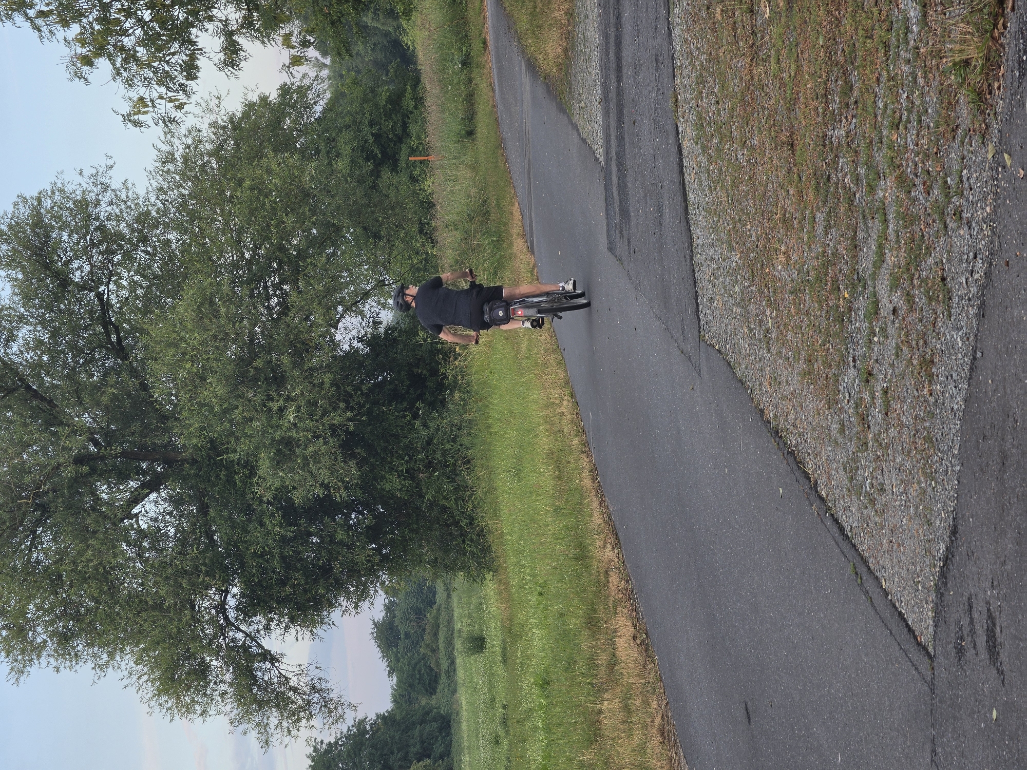 A lone person wearing a black t-shirt, black shorts, a black helmet, and white sneakers is cycling on a paved path in Kelsterbach, Germany. The cyclist is moving away from the camera on a bicycle with a black frame and a small black bag attached to the rear. The path is asphalt and has a gravel shoulder. To the left of the path is a grassy field, and to the right, the path curves and leads into a wooded area with large trees. The sky is overcast with light clouds. The overall scene depicts a quiet outdoor activity in a natural setting.