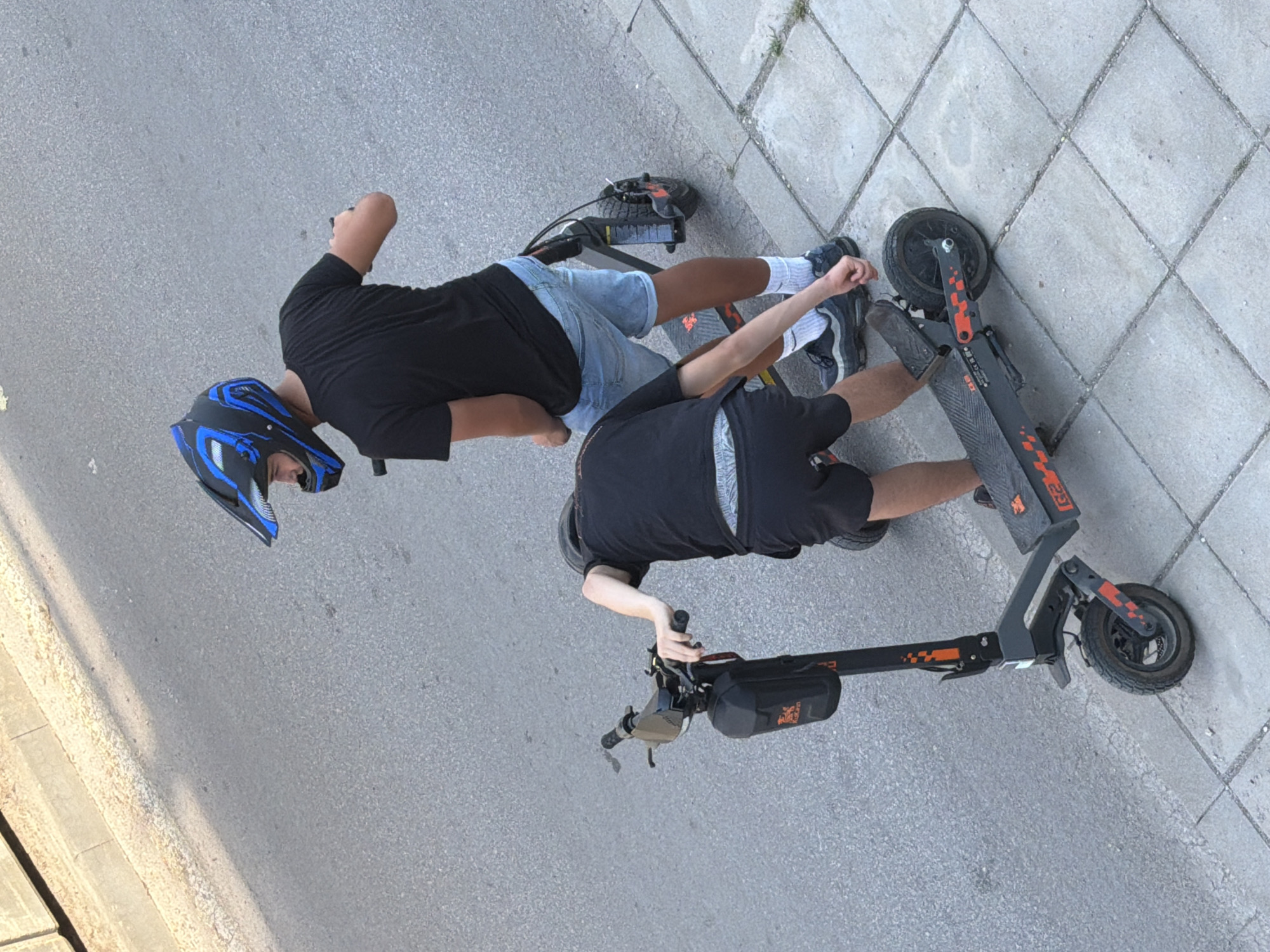 Two individuals are standing next to electric scooters on a sidewalk bordering a paved road. The individual in the foreground, facing away from the camera, is wearing black shorts, a black t-shirt, and a helmet. They are leaning over one of the scooters, with their right hand on the handlebar. The scooter is black with orange accents and the model number "G2" is visible.
The individual in the background, also facing away from the camera, is wearing denim shorts, a black t-shirt, and a blue and black motocross-style helmet. They are standing next to a second electric scooter, which appears similar to the first. Both individuals are wearing athletic shoes and white socks.
The environment consists of a grey asphalt road and a light-colored tiled sidewalk. The lighting suggests it is daytime, with sunlight casting shadows. The scene appears to be in an urban setting.
