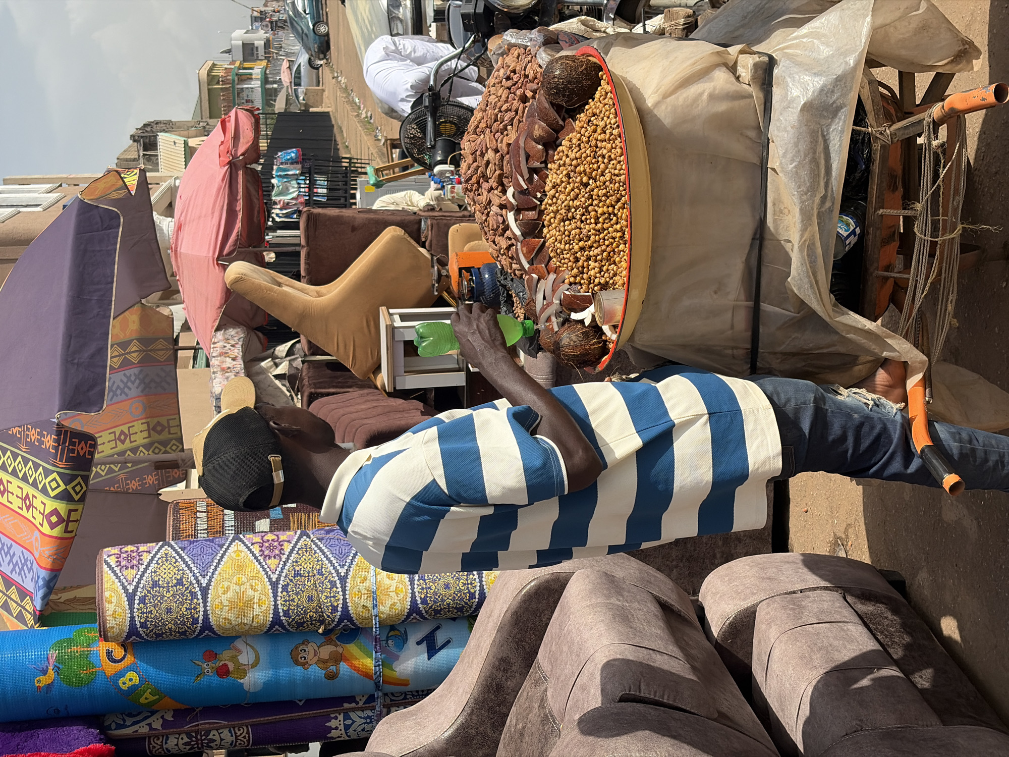 The image captures a street scene in the city of Jos, Nigeria, where a man wearing a blue and white striped polo shirt and a black baseball cap stands next to a large tray of food items, likely nuts and coconuts, displayed on a cart. He is holding a green plastic bottle. Behind him, rolled-up mats and fabrics are stacked, suggesting a market stall. In the background, more market stalls, buildings, parked cars, and people are visible along a dusty street under an overcast sky. The overall scene depicts a typical day in a bustling Nigerian marketplace.