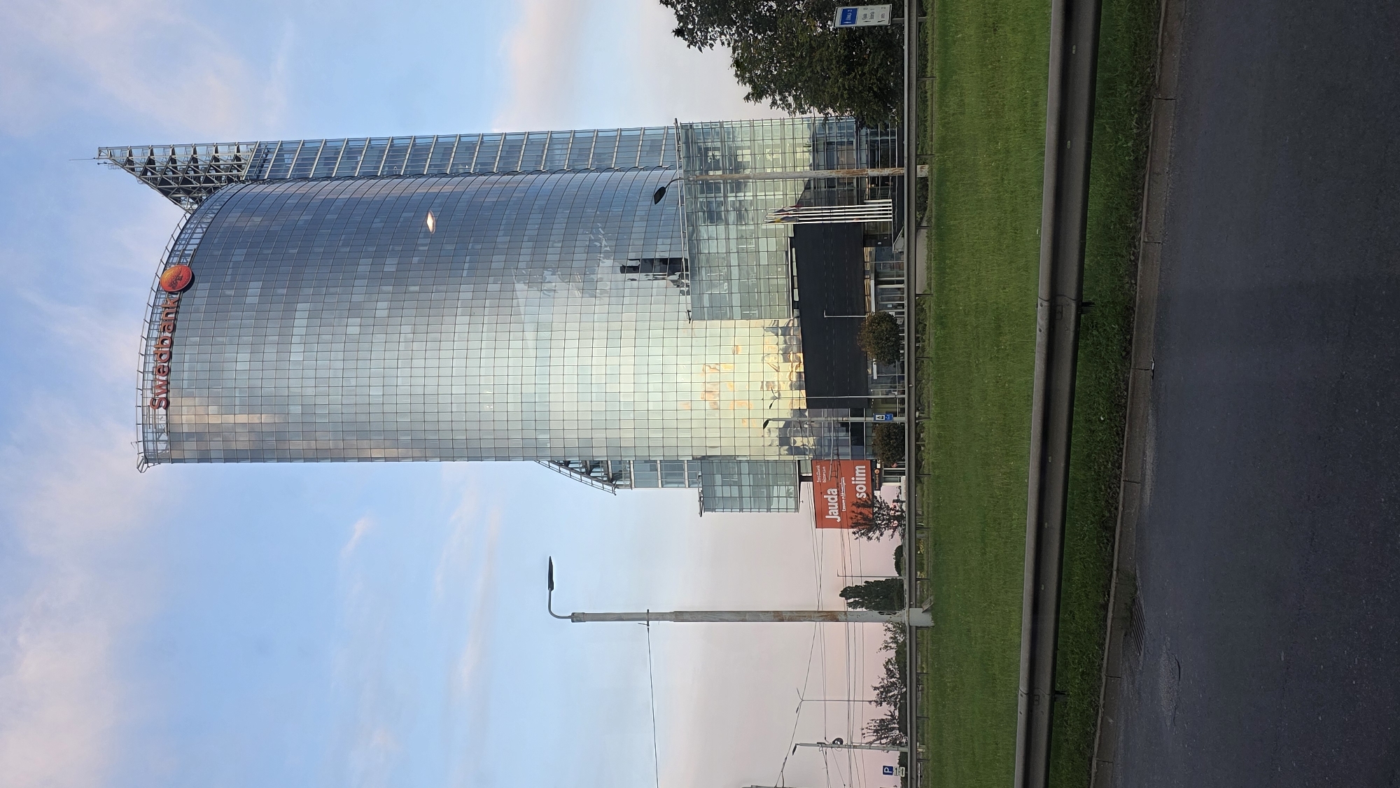The image depicts the Swedbank headquarters building in Riga, Latvia, at approximately dusk. The modern skyscraper, characterized by its curved glass facade, reflects the soft hues of the twilight sky and surrounding environment. A prominent "Swedbank" logo is visible at the top of the building. In the foreground, a grassy median separates the road from the building. Several streetlights and traffic signals are present along the roadside. A billboard advertising "Jauda.solim" is also visible near the base of the building. The sky exhibits a gradient of blue and pink, indicating the transition from day to night. The scene is devoid of any visible people or vehicles in immediate proximity to the building.