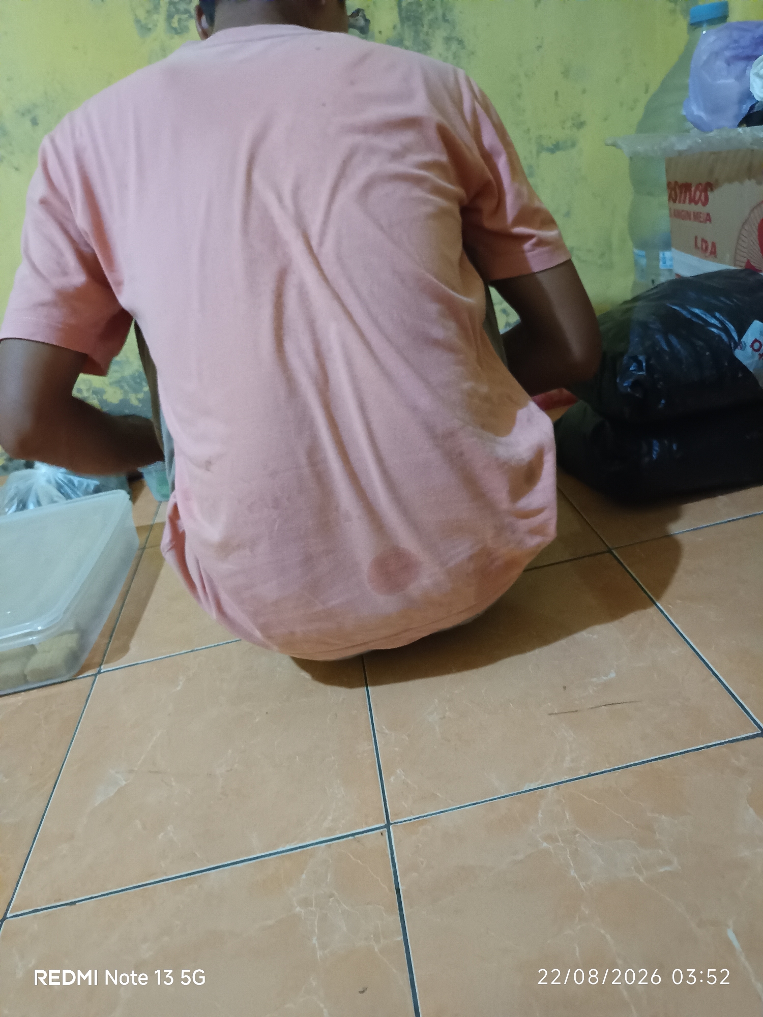 The image shows a person from behind, wearing a light pink t-shirt, seated on a tiled floor. The t-shirt appears to be damp in patches. To the right of the person are two black plastic bags stacked on top of each other, and behind them, a cardboard box with the brand name "OSMOS" visible. A blue plastic bottle is also present. To the left of the person, a clear plastic container holding what appear to be food items is on the floor. The background wall is a textured yellow. The floor tiles are a light brown with darker grout lines. The timestamp in the bottom right corner indicates the image was taken on August 22, 2026, at 03:52. The text "REDMI Note 13 5G" appears in the bottom left corner, indicating the device used to take the photo. The scene is indoors, likely in a residential setting in Talaga Sari, Indonesia.