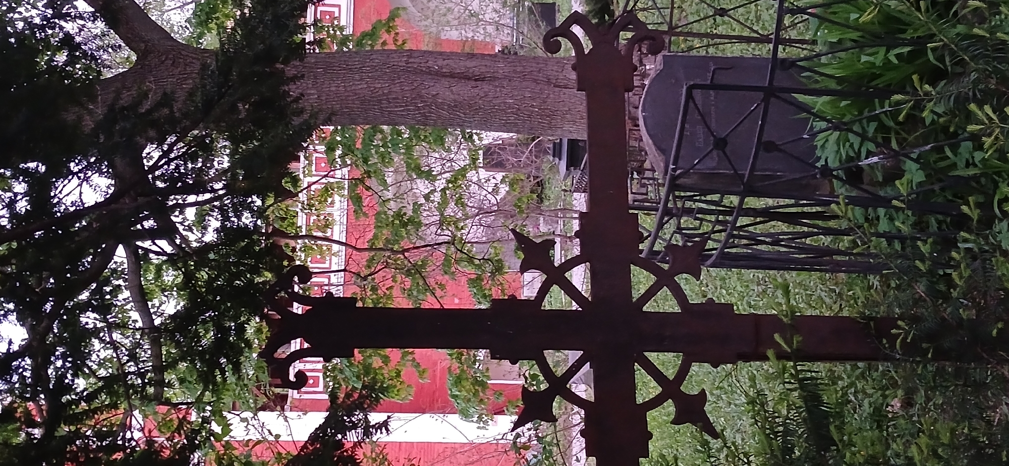 The image depicts a weathered iron cross in a cemetery in Moscow, Russia. The cross has an intricate design with decorative flourishes. In the background, a red brick building with white trim is visible through the trees. Other graves and wrought-iron fencing are scattered in the background. The scene is partially obscured by vegetation, suggesting a neglected or overgrown area within the cemetery.