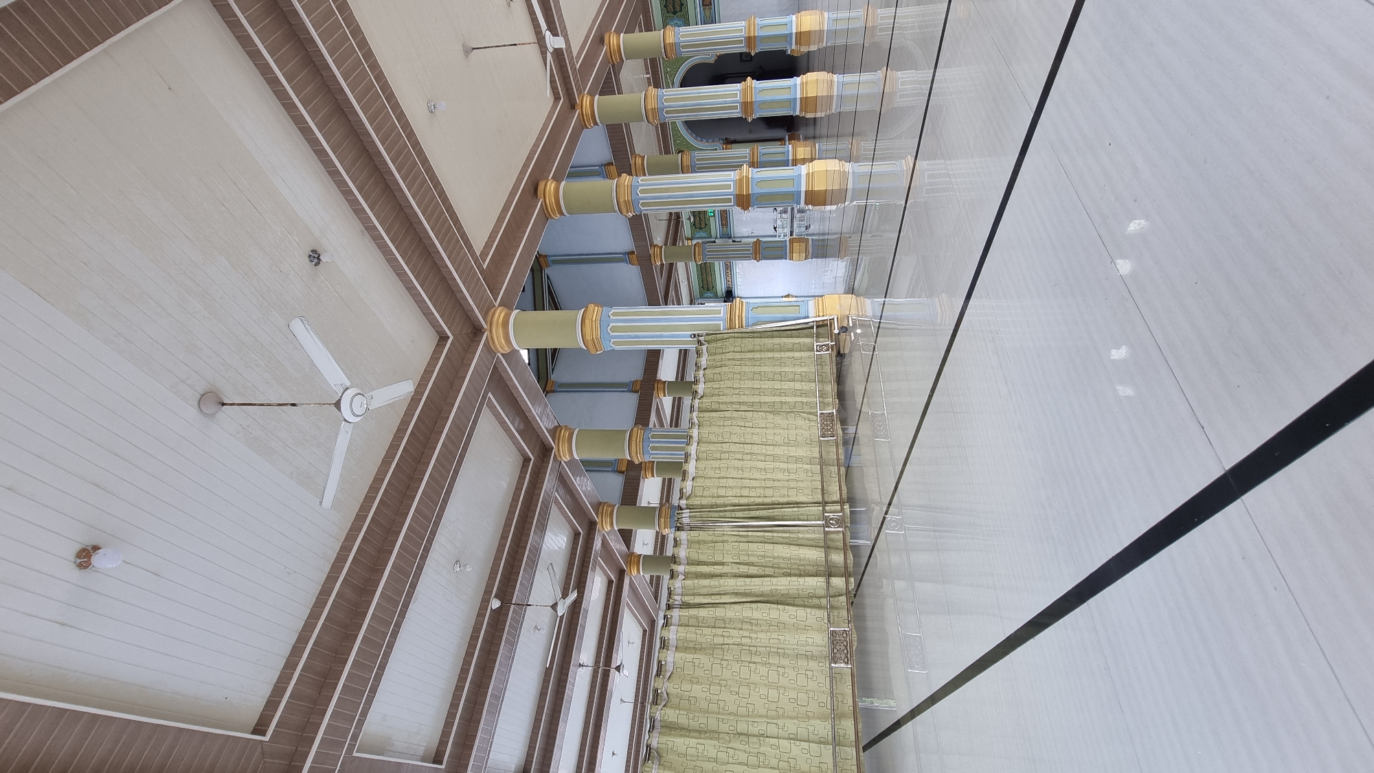 The image depicts the interior of a large, empty hall, likely a mosque, in Meunasahlhok, Indonesia. The hall features a high, coffered ceiling with recessed lighting and several white ceiling fans. Large, decorative pillars with yellow, blue, and green detailing support the ceiling. A long, light green curtain with a subtle geometric pattern stretches across the middle of the hall, supported by metal railings. The floor is highly polished white tile with prominent black lines, reflecting the ceiling and pillars. The scene is static, with no people or discernible actions. The environment appears clean and well-maintained, suggesting a place of worship or public gathering that is currently unoccupied.
