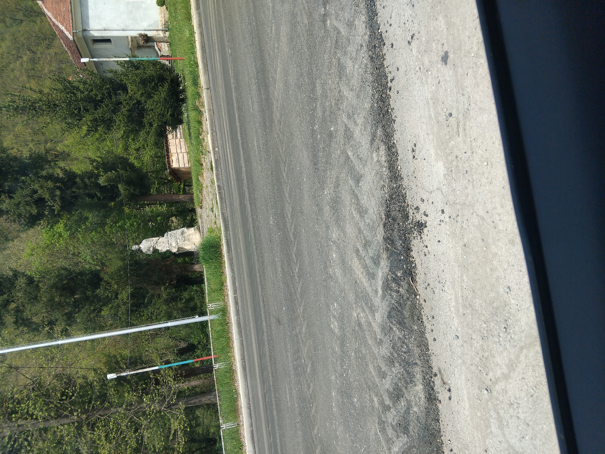 The image displays an outdoor scene, likely captured from a vehicle. A paved asphalt road, with tire marks faintly visible, stretches from the foreground towards the background, dominating the central and right portions of the frame. To the immediate right of the road is a lighter-colored concrete shoulder or curb, bordered by a dark, indeterminate object (possibly part of a car's interior or frame) on the far right, casting a shadow onto the concrete.
On the left side of the road, a vibrant green grass verge runs parallel to the asphalt. Beyond the grass, a dense area of lush green trees, comprising both coniferous and deciduous types, forms a significant part of the landscape. Among these trees, a light-colored building with a distinct reddish-brown tiled roof is partially visible in the mid-ground. Closer to the foreground on the left, a weathered stone statue, depicting a human figure, stands erect. Two utility poles are prominent features on the left: a tall, slender silver-grey pole and a shorter white pole adorned with notable red and blue bands, both supporting overhead wires that extend across the scene. A small, rustic wooden structure or bench can also be discerned nestled within the trees.
The scene is brightly lit by natural sunlight, indicating a clear, daytime setting. There are no visible people or discernible text within the image. The overall environment suggests a semi-rural or exurban area, characteristic of the specified location of Velchevo, Bulgaria.