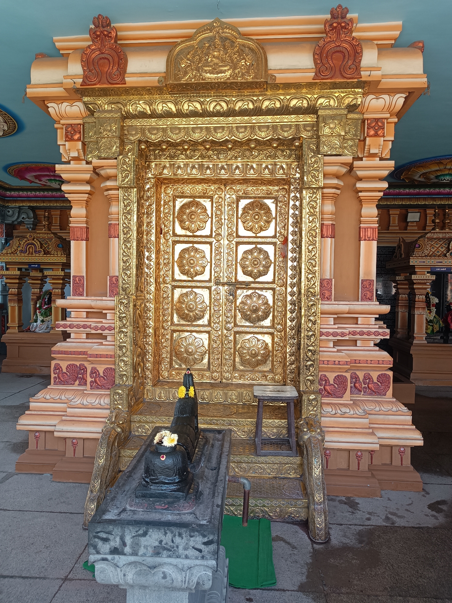 The image depicts a highly ornate, golden doorway in what appears to be a Hindu temple. The doorway is rectangular and features intricate floral designs on its surface. It is flanked by peach-colored pillars adorned with carved details and topped with reddish-brown decorative elements. A structure resembling a stepped platform leads up to the doorway, with decorative carvings along its edges. In front of the doorway, on a gray stone base, sits a black Shiva Lingam adorned with flowers. A small wooden stool rests to the side of the Lingam. A green cloth is situated at the base of the stairs. The overall environment suggests an interior space of a temple, given the architecture and religious iconography. Based on the prompt, the location is Ayanambakkam, India.