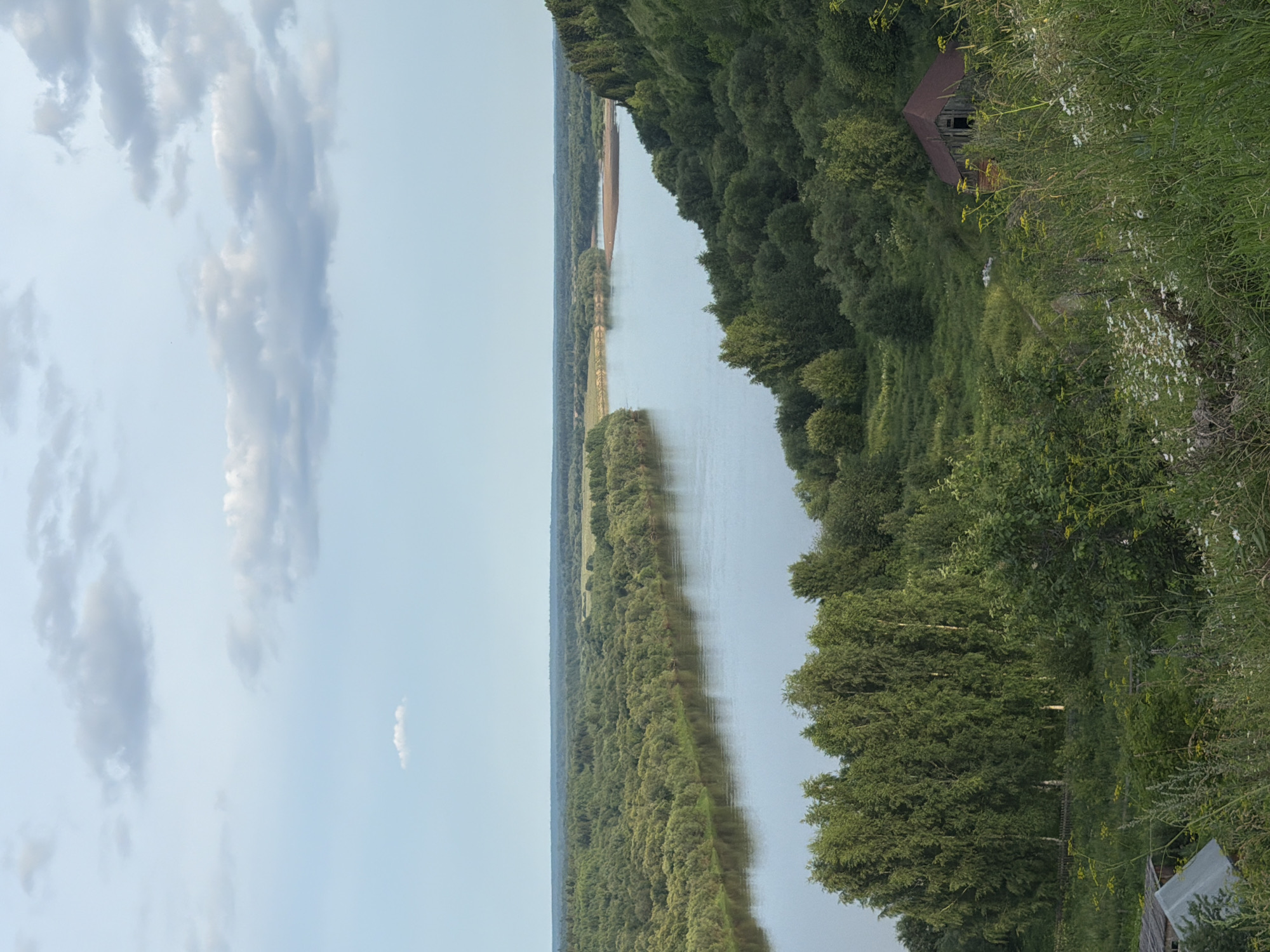 This image depicts a scenic view of a wide river flowing through a lush, green, forested landscape. The river curves gently, with its water reflecting the pale blue sky and scattered clouds. The foreground features a grassy hillside with wildflowers and some trees. In the mid-ground, a dense forest covers the rolling hills on either side of the river. On the right side of the frame, nestled within the trees, is a rustic, abandoned wooden building with a dark, reddish-brown roof. In the distance, the river opens up to a wider expanse, with more trees and a flat plain stretching to the horizon. The overall impression is one of natural beauty and tranquility, with a touch of rural abandonment. The location is the city of Cherdyn, Russia, specifically a view overlooking the Kama River.