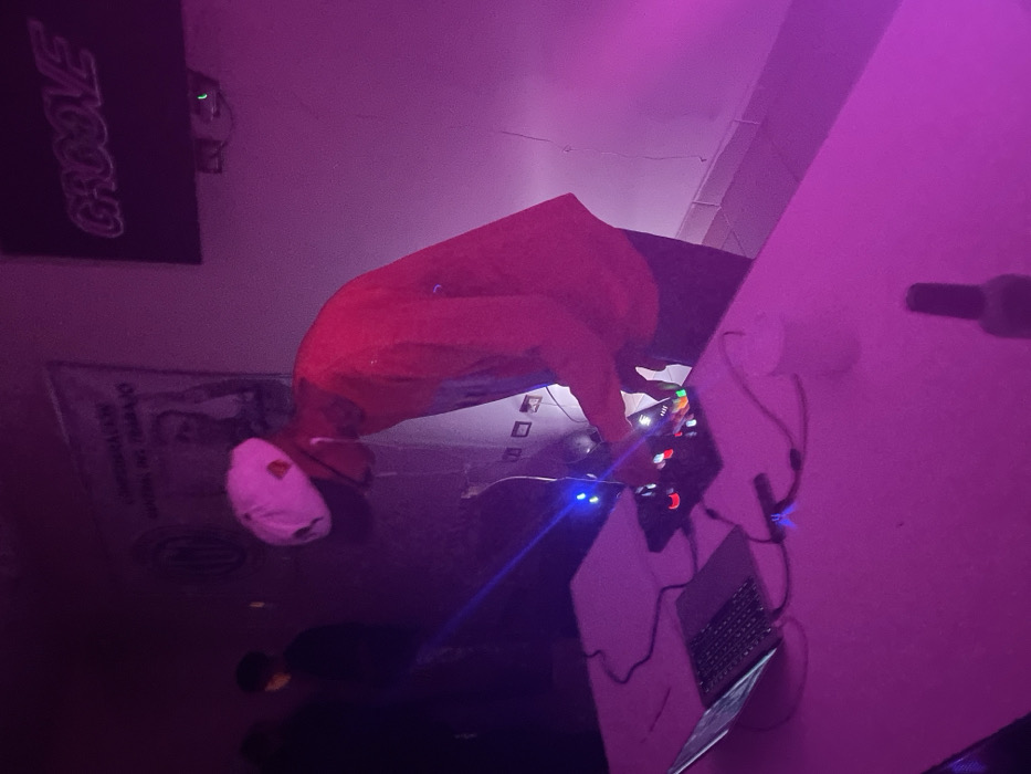 The image depicts an indoor scene in Ushuaia, Argentina, characterized by dim, purple-pink ambient lighting. A male individual, positioned slightly right of center, wears a white baseball cap reversed, dark glasses, a red long-sleeved shirt, and dark trousers. He is leaning forward, actively manipulating a black DJ controller that emits blue light, positioned on a white table. A silver laptop, open, rests to the left of the controller, with various cables connecting the devices. A white cup is visible near the controller, and a dark, slender bottle stands in the immediate foreground.
In the background, against a light-colored wall, a banner is affixed, partially legible with the text "CONFEDERACION GENERAL DEL TRABAJO" and a circular emblem, alongside an obscured portrait. To the right of the banner, a dark rectangular sign displays the illuminated word "GROOVE." Another person, largely silhouetted in a dark top, is partially visible in the far left background. The wall features visible power outlets and minor surface imperfections.