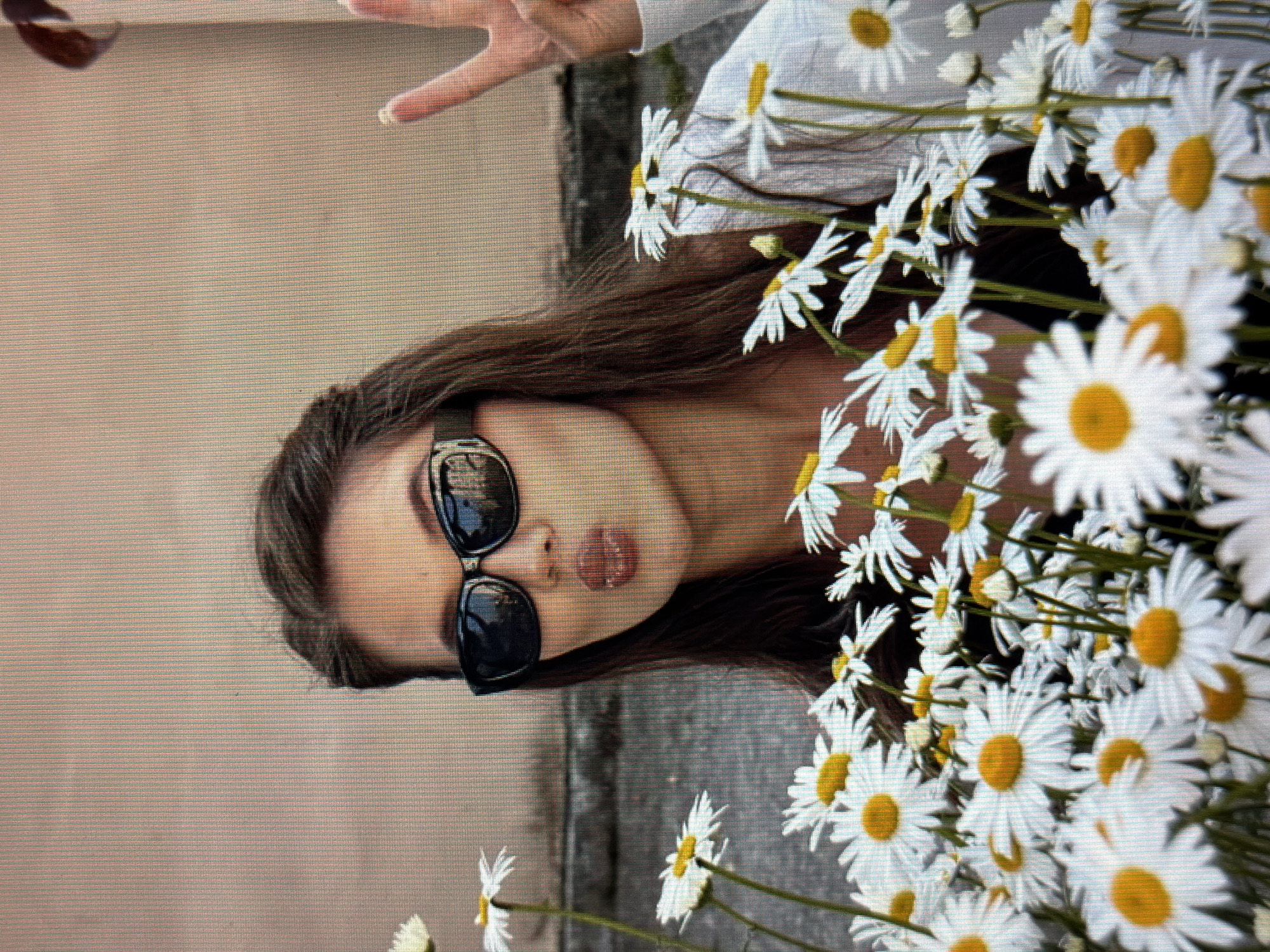 A young woman with brown hair, wearing black sunglasses and a white shirt, poses for a photo in front of a wall with a bouquet of daisies. She is puckering her lips and holding up two fingers in a peace sign. The background is a light pink wall with a darker textured area near the ground. The image appears to be taken outdoors in a city setting. No specific details about the city of Sirius, Russia, are visible in the image.