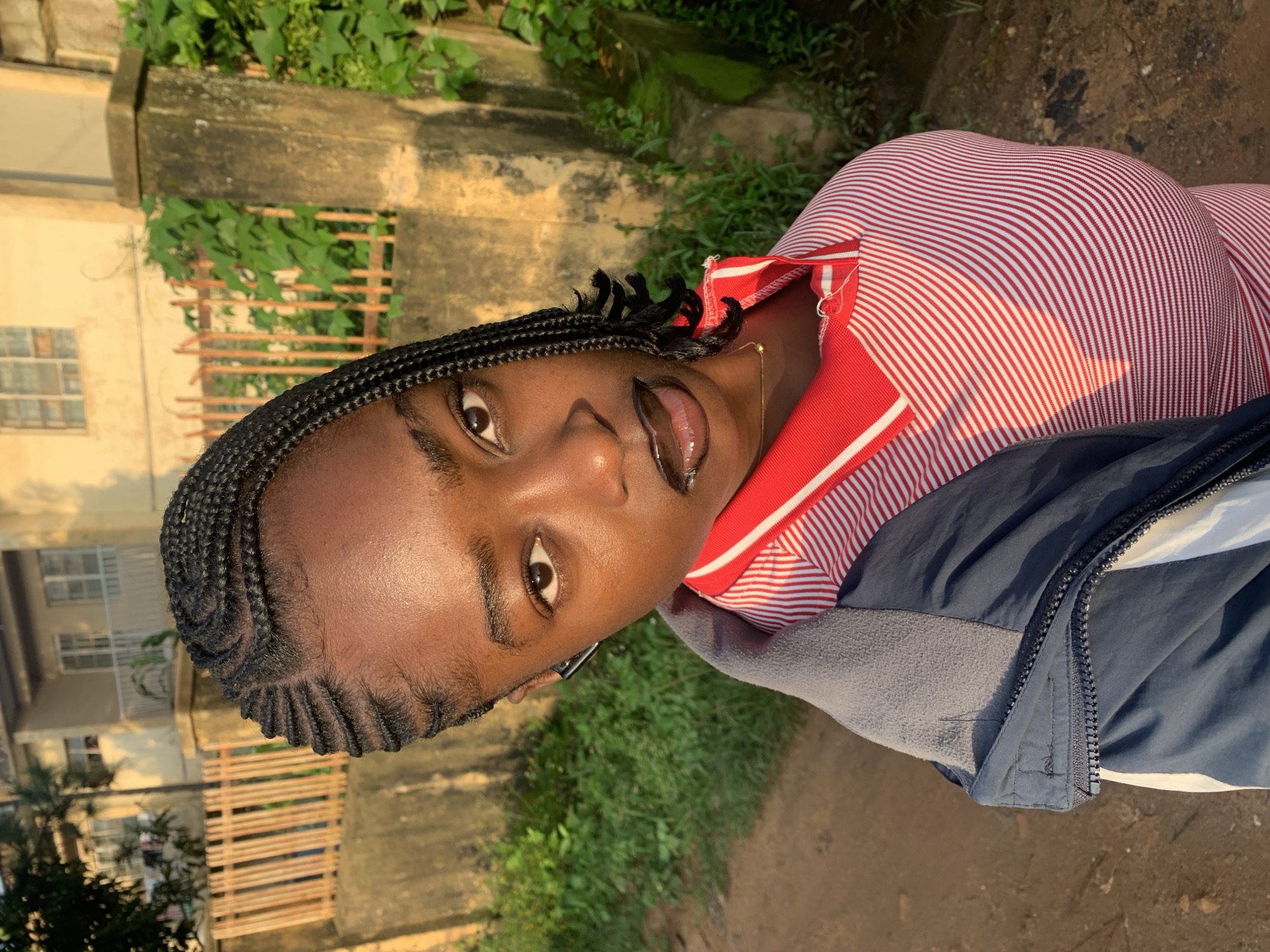 A young Black female is shown in a medium close-up, angled to her right, outdoors in what appears to be a residential area. She has dark skin and her hair is styled in neat, dark braids that are pulled back from her forehead. She is wearing a red and white striped polo shirt with a grey and navy jacket layered over it. The jacket is zipped up to her neck. Her eyes are brown, and she is wearing dark lipstick. A small gold chain is visible around her neck. In the background, there is a textured wall, a wooden fence, lush green vegetation, and a building with balconies and windows. The lighting suggests it is late afternoon or early morning, casting long shadows and highlighting the textures of the environment. The location is identified as Rafin Par, Nigeria.