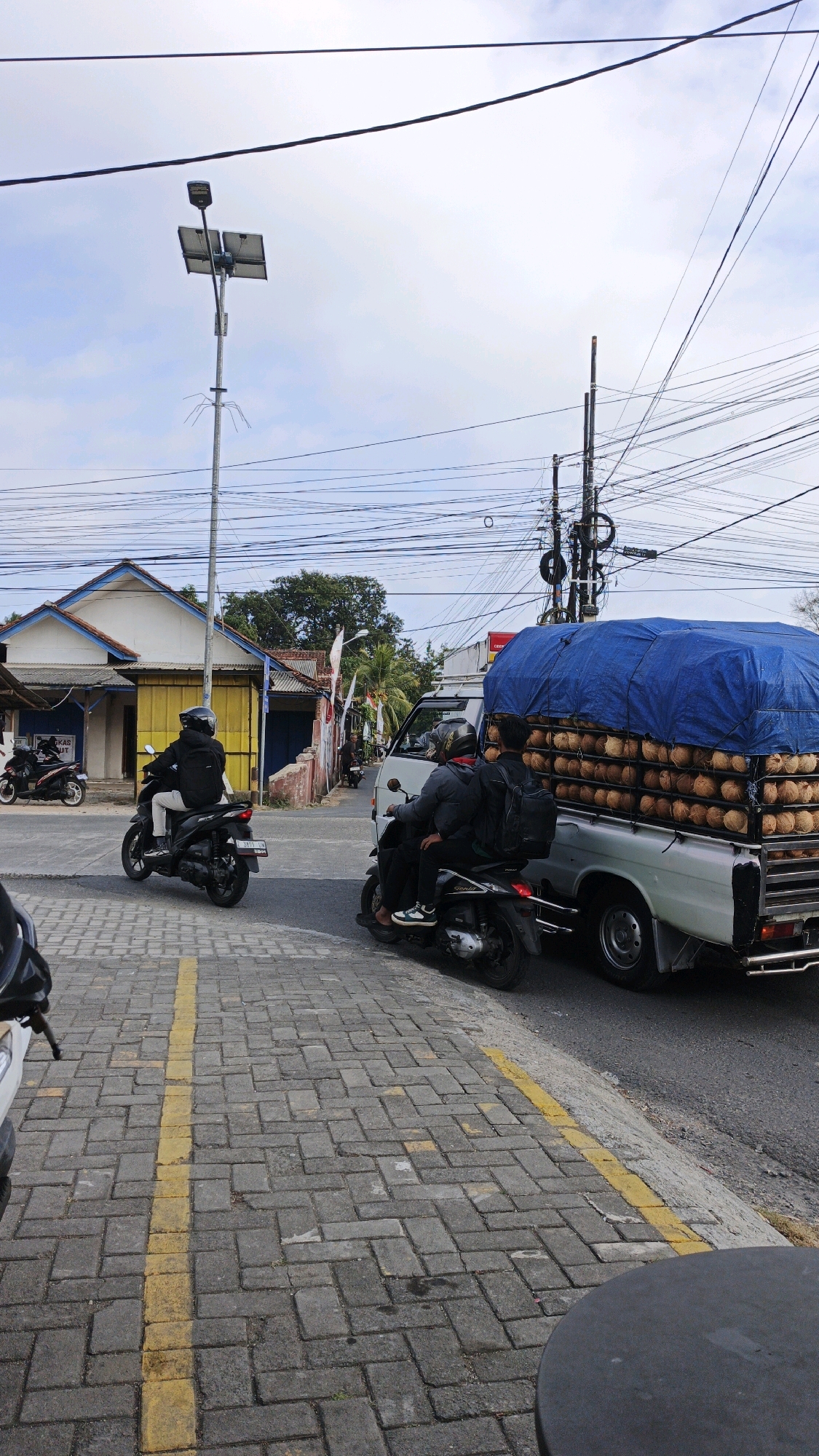 The image shows a street scene in Cibenda, Indonesia. In the foreground, a paved walkway with a yellow line runs along the side of a road. On the road, two motorcycles are visible. One motorcycle, a black scooter, is being ridden by a person wearing a black jacket and helmet, with a black backpack. This motorcycle is positioned to the left of the frame. To the right, another black scooter carries two people, both wearing jackets and helmets, also with backpacks. They are positioned behind a white pickup truck.
The pickup truck is loaded with a large quantity of coconuts, which are covered by a blue tarp. The truck is facing towards the right of the frame. In the background, there are buildings with yellow and white walls and blue trim, as well as palm trees and a tangled mess of power lines and utility poles against a cloudy sky. A solar-powered street light is also visible. The overall impression is of a busy street in a tropical urban environment.