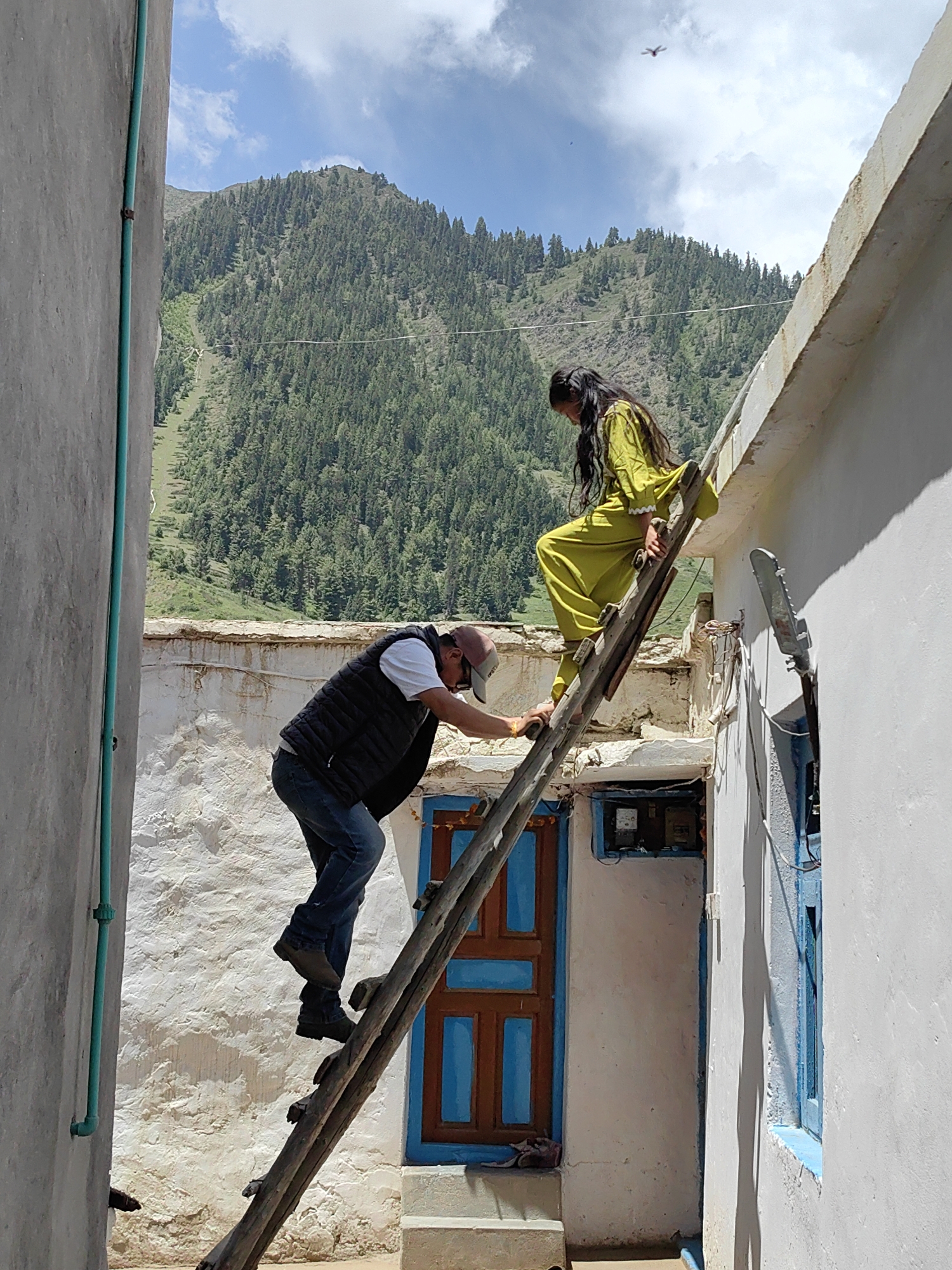 A man wearing a vest, t-shirt, jeans, and a cap is climbing a wooden ladder. A young woman in a lime green outfit is standing on the roof next to the ladder, holding onto it. The scene takes place outdoors, with a stucco wall on the left, a building with a blue-trimmed door and window in the background, and a mountainous, tree-covered landscape visible beyond the roof. A bright blue sky with clouds and a distant airplane is overhead. The overall impression is one of daily life activity in a rural or mountainous region of Kishorl, India.