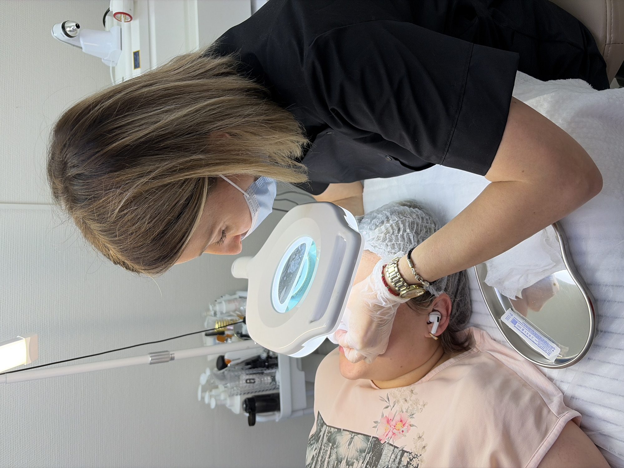 The image shows a woman in a black shirt and surgical mask performing a cosmetic procedure on a patient lying on a white treatment bed. The patient has a white mesh cap on and is wearing AirPods. The cosmetic provider uses a magnifying lamp to examine the patient's face. A metal tray holding swabs sits next to the patient. Various medical devices are visible in the background. The location appears to be a clinic or spa setting in an indoor environment in the city of Unknown, Unknown.