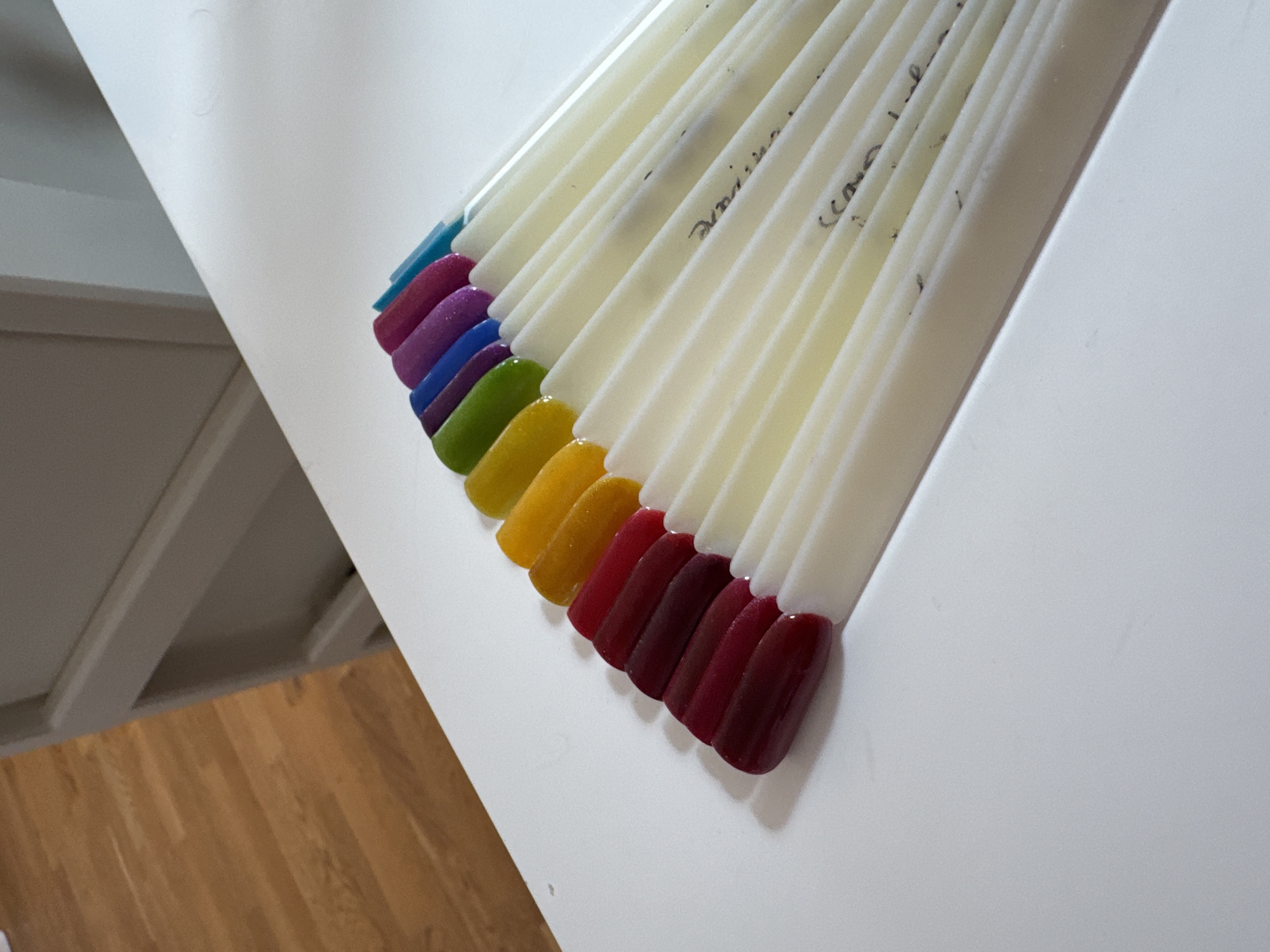 A set of nail polish swatches is fanned out on a white surface. The swatches are arranged in a rainbow gradient, starting with deep red, transitioning to lighter reds, then yellow, green, blue, purple, and finally a bright blue at the end of the fan. The swatches are attached to white plastic sticks, and some of the sticks have handwritten labels in black ink. The background consists of a light-colored wooden floor and a portion of a white cabinet.