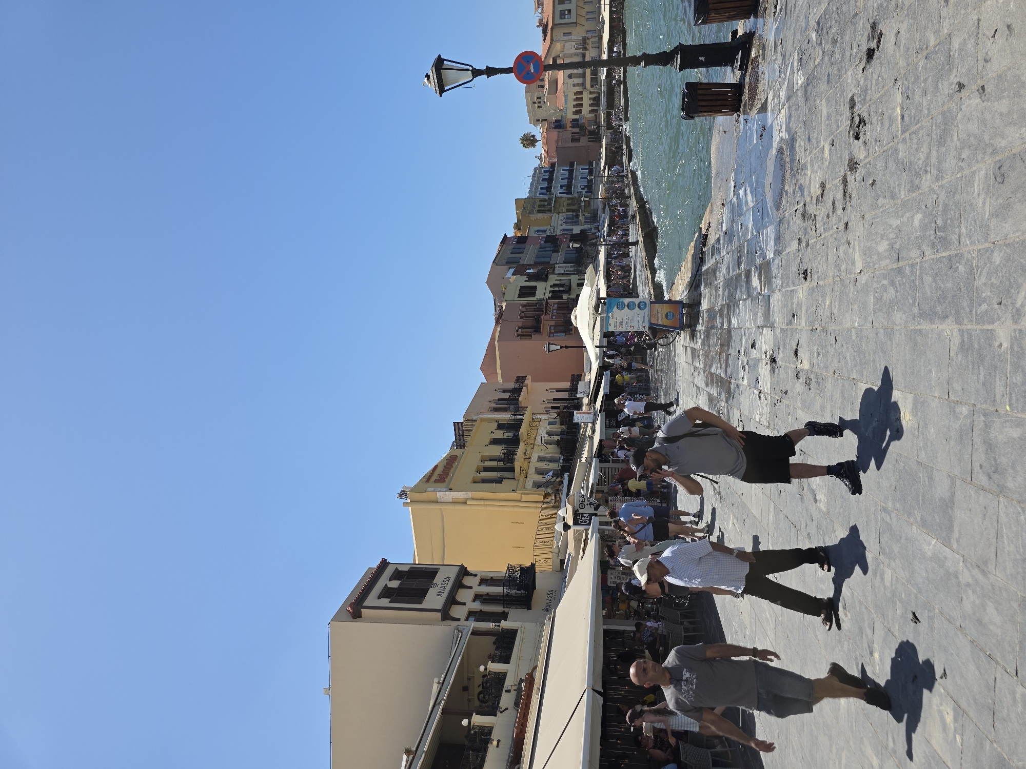 The image depicts a busy waterfront promenade in the city of Chania, Greece, likely the Venetian harbor. A line of colorful buildings with balconies and awnings runs along the left side of the scene, housing shops and restaurants. The promenade is paved with large stone slabs and is crowded with pedestrians. In the foreground, three men are walking. The man on the left wears a gray t-shirt and denim shorts. The man in the middle wears a checkered shirt and dark pants. The man on the right wears a gray polo shirt, black shorts, and a backpack. The sea is on the right, with choppy water under a clear blue sky. A lamppost with a "no stopping" sign is visible near the water's edge. Several trash cans are placed along the promenade. The overall impression is a vibrant, sunny day at a popular tourist destination.