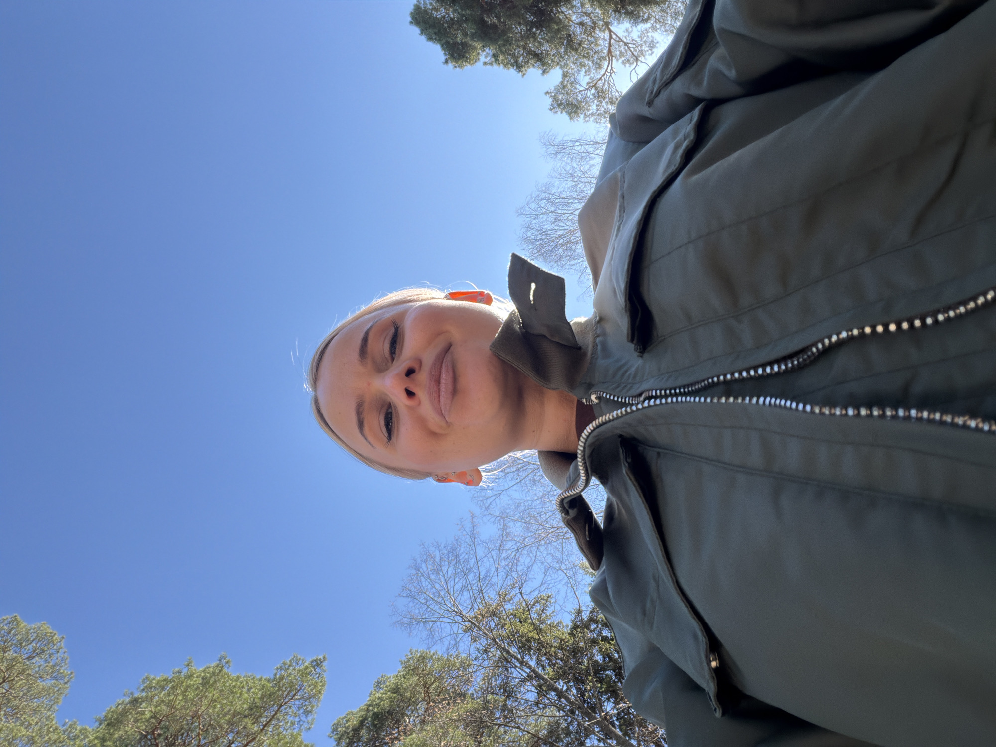 A low-angle selfie taken outdoors depicts a young woman with blonde hair and fair skin. She is wearing a green, zippered jacket. The background shows a clear blue sky and treetops, suggesting a wooded area or park. There is no specific evidence in the image to confirm the location is Sarapul, Russia.
