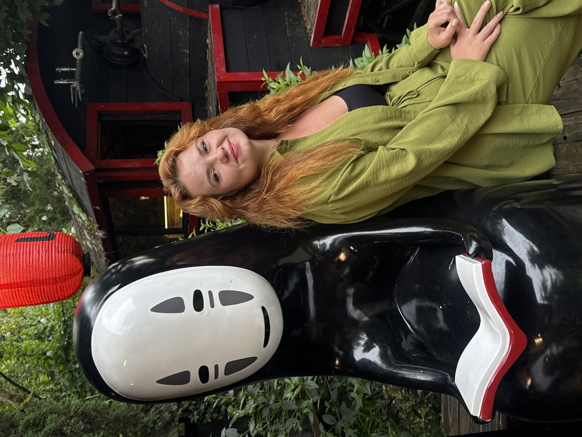 In Cam Ly - Đà Lạt, Vietnam, a young woman with auburn hair and freckles is pictured sitting next to a statue of the character No-Face from Spirited Away. She is wearing a green collared shirt and matching pants, with a black top underneath. Her right arm is resting on her lap, and her left arm is on the statue. The No-Face statue is black and white with a white face and black eyes and mouth. It is holding an open book with red covers. Behind them, a red lantern hangs from a tree, and a dark-colored structure with red trim resembling a train car is visible. Lush green foliage surrounds the scene, suggesting an outdoor setting.