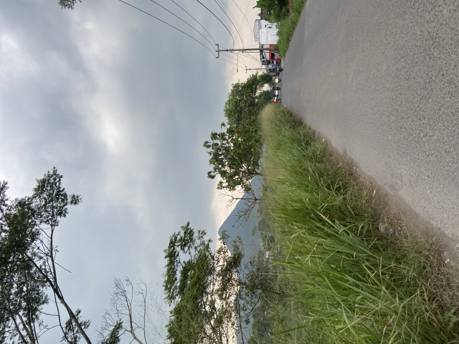 The image shows a view of a paved road in Kecamatan Margaasih, Indonesia, looking down the road towards a small town. The road is bordered by tall grass and trees on the left, and a few buildings are visible on the right. Several motorcycles are driving down the road. There are power lines overhead. In the distance, a mountain can be seen through the overcast sky.
