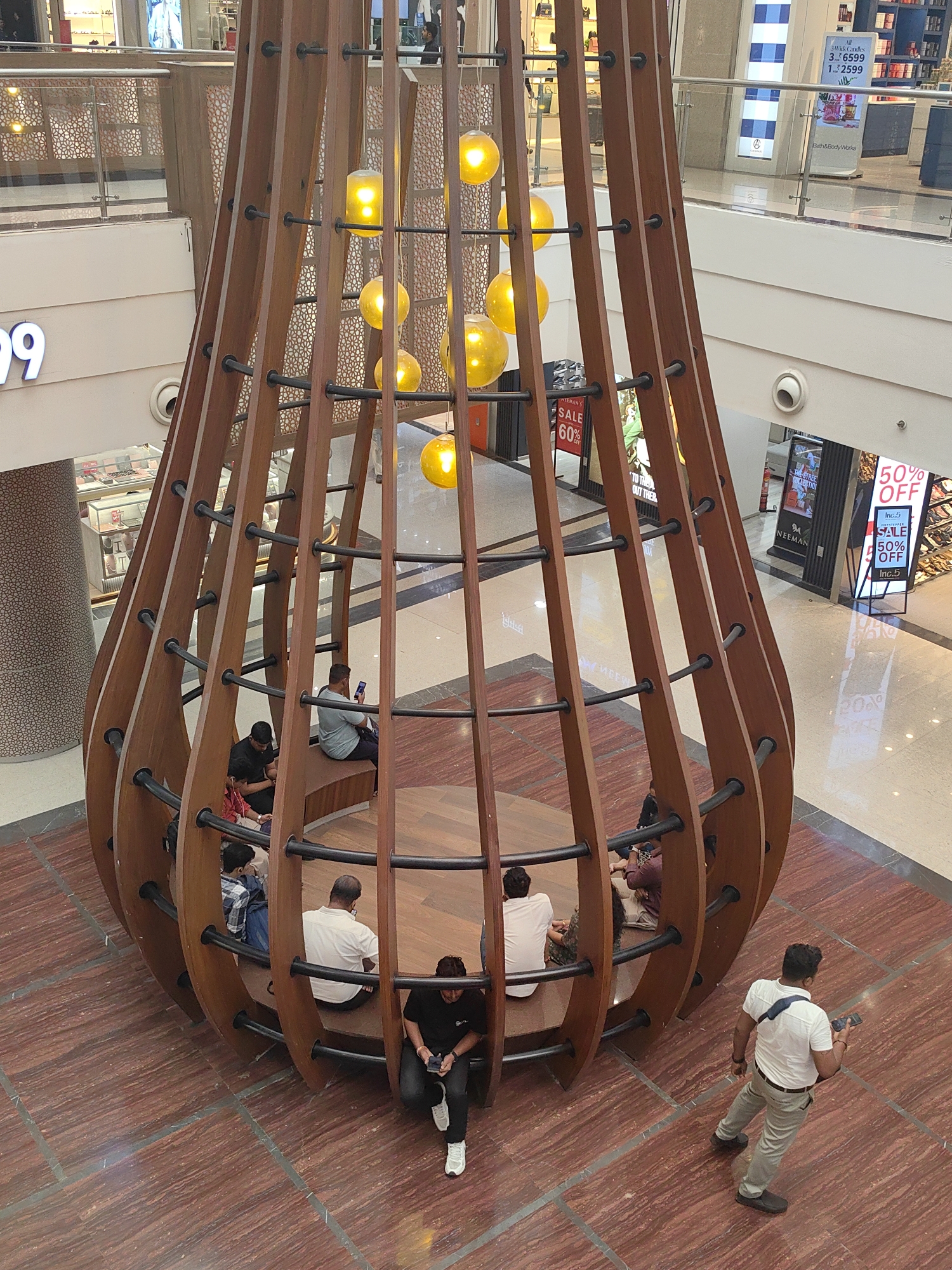 The image captures an interior view of a modern shopping mall, likely located in Mumbai, India, given the presence of a prominent architectural installation. Dominating the foreground is a large, wooden, sculptural structure resembling an upside-down bell or a large hanging pod, with a lattice-like exterior made of curved wooden planks and black metal supports. Several warm yellow pendant lights hang from the ceiling within this structure, illuminating the space.
Inside the wooden structure, there is a circular wooden seating area. Several individuals are seated on this bench, engaged in various activities. One person is looking at their phone, another is sitting with their legs crossed, and others are seated more passively. The flooring inside the structure is also wooden.
Outside the structure, the mall's polished red and beige tiled floor is visible. To the right of the frame, a man in a white t-shirt and light-colored trousers is walking away from the camera, holding a mobile phone in his right hand. He is wearing a dark blue strap across his chest.
In the background, the multi-level architecture of the mall is apparent, with glass railings and shops visible on different floors. Signage is present, including a large blue sign with white text that reads "99" and other commercial advertisements on display. The overall environment suggests a public, commercial space with a focus on modern design and consumer activity.