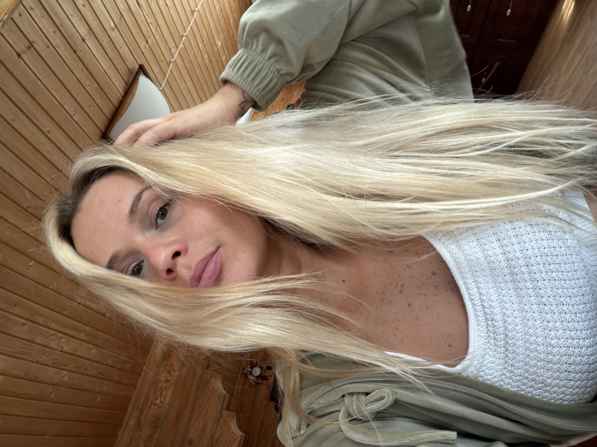 A selfie angled downwards showing a fair-skinned woman with long blonde hair. She is wearing a white knitted top and a light green jacket. Her left hand is visible near her head. The background is a wood-paneled wall and ceiling. A small window is visible on the upper left. Location cannot be determined from the image.