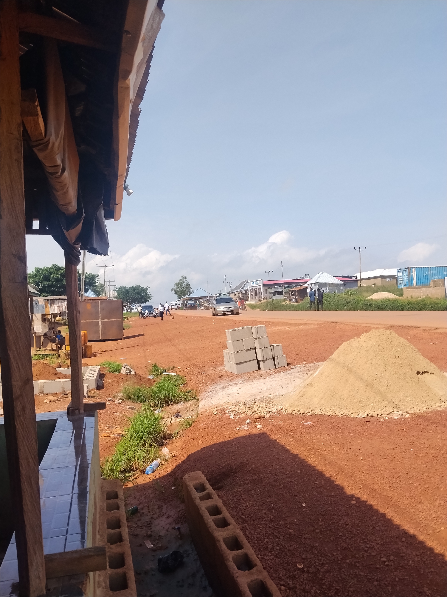 The image shows a street scene in Durumi, Nigeria, on a clear day. The foreground features construction materials, including piles of sand and stacks of concrete blocks, along with a partially built structure with tiled surfaces and wooden supports. In the middle ground, a red dirt road runs horizontally, with several vehicles and individuals present. To the left, there are some metal structures and what appear to be stalls or shops. Further down the road, buildings line the street, some with corrugated metal roofs and others with more permanent structures. Several people are visible, some standing and others walking. The sky is a pale blue with scattered white clouds. The overall impression is that of an active area with ongoing construction and daily life unfolding along a roadway.