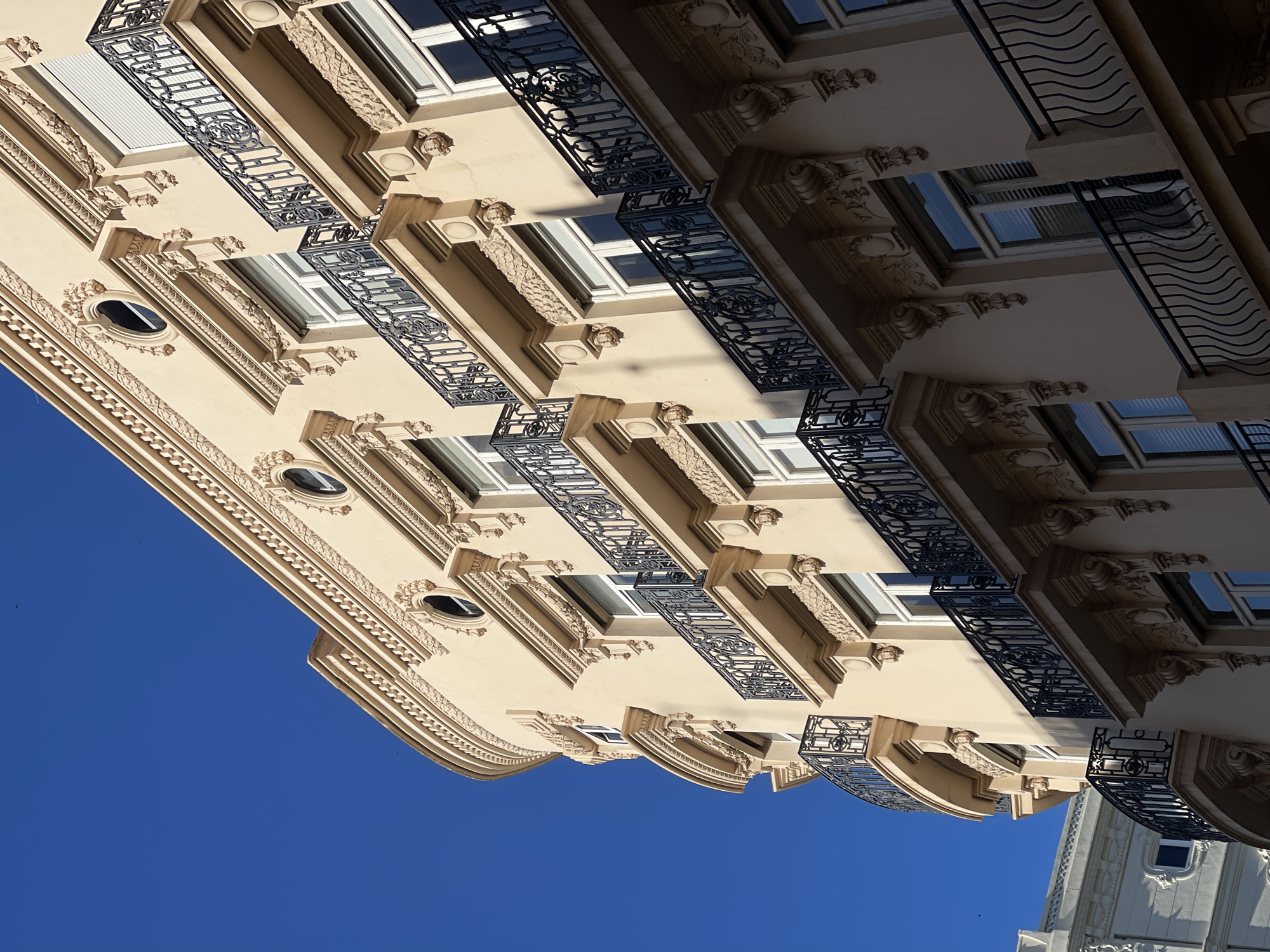 The image shows a low-angle view of a section of a building in València, Spain, likely a residential or commercial structure, showcasing its architectural design and details. The building is several stories tall with a light beige facade and ornate detailing, including decorative moldings, window surrounds, and sculpted embellishments. Each level features windows and balconies. Some balconies have intricate black iron railings with decorative patterns. The sky is bright blue and clear. The overall impression is of a well-maintained, classic European architectural style, with focus on the upper part of the building.