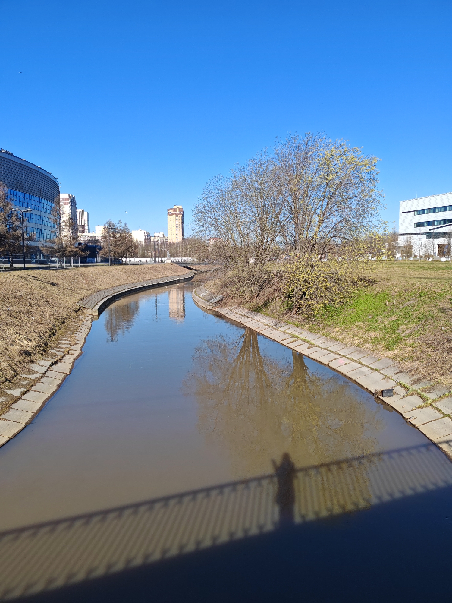 The image depicts a canal in Saint Petersburg, Russia, under a clear blue sky. The canal, lined with concrete blocks, contains murky water reflecting the bare trees on its banks. On the left, modern buildings are visible, while the right bank transitions into a grassy area. The shadow of a bridge railing and a person are cast onto the water in the foreground.