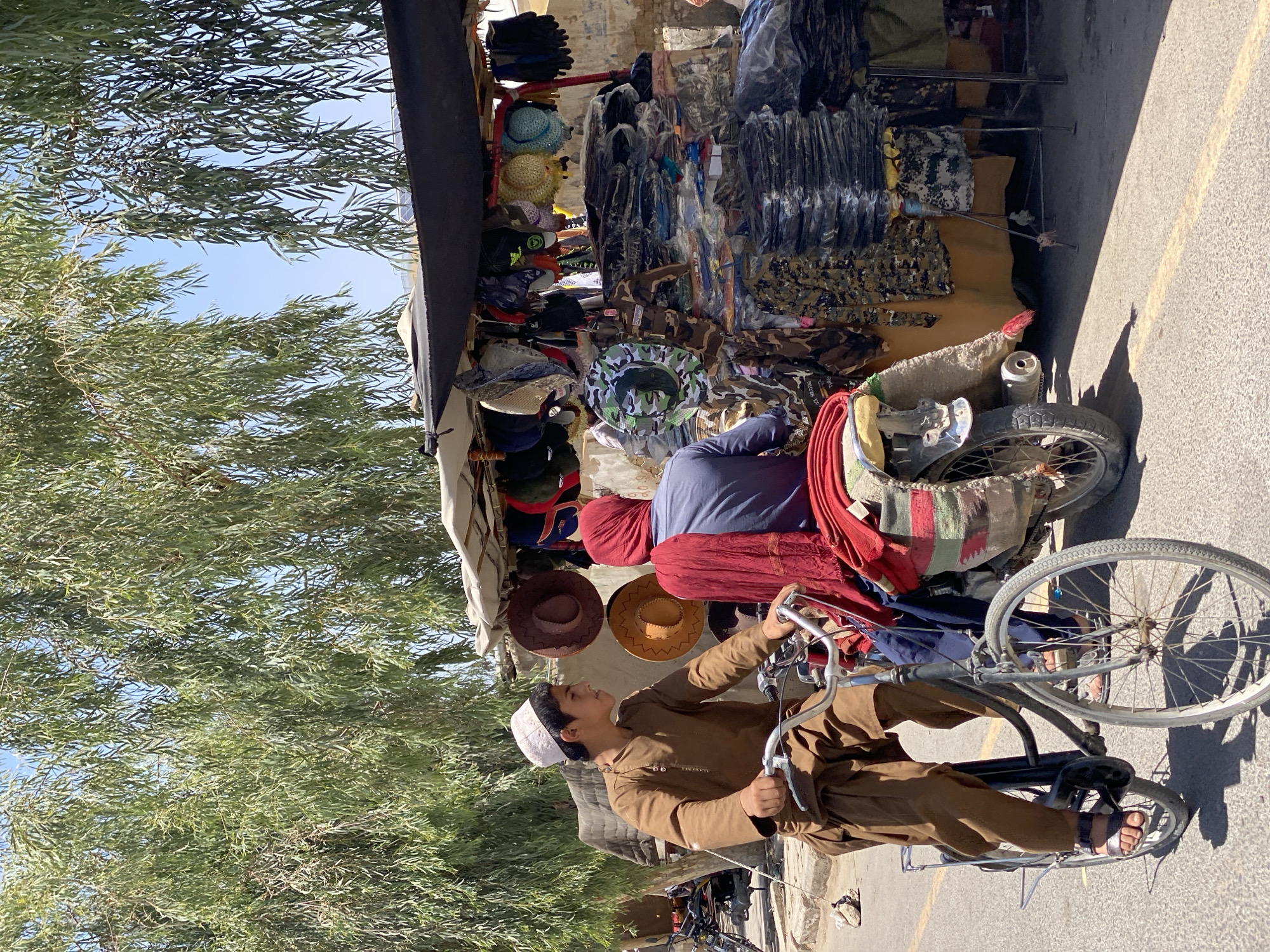 In Kandahar, Afghanistan, a young boy wearing a traditional brown outfit and a white cap rides a bicycle on a paved road. He is positioned in the foreground, with his profile facing right. Behind him, a person wearing a red headscarf and a blue shirt sits on the back of a motorcycle, also facing right. The motorcycle is parked alongside a street vendor's stall, which is laden with a variety of goods, including hats, scarves, and folded clothing. The stall is covered by a black awning, and displays items on racks and shelves. The background features a large tree with drooping branches, its leaves creating a dappled shade on the scene, and a clear blue sky. The environment appears to be a bustling marketplace on a sunny day.