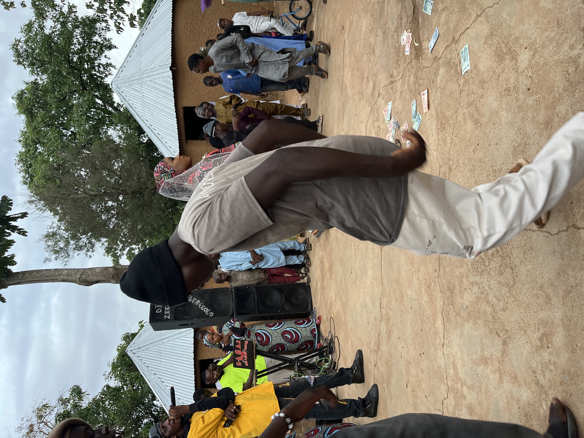 An outdoor gathering takes place in Jos, Nigeria, on a cloudy day. In the foreground, a young man in a gray t-shirt and white pants is dancing. His body is angled away from the camera, and he is in mid-motion with his arms bent. Scattered on the ground around him are several Nigerian Naira banknotes in denominations of 10 and 20. Behind him, a group of people are gathered. To the left, a man in a yellow t-shirt and black pants holds a microphone, appearing to be speaking or singing. A DJ speaker is positioned behind him. Several other individuals are visible, including a woman in a colorful headscarf and dress standing near the speaker, and men in traditional attire. Two small, mud-brick buildings with corrugated metal roofs are visible in the background, along with trees and vegetation. A bicycle is parked to the right of the frame. The ground is a mix of dirt and cracked concrete.