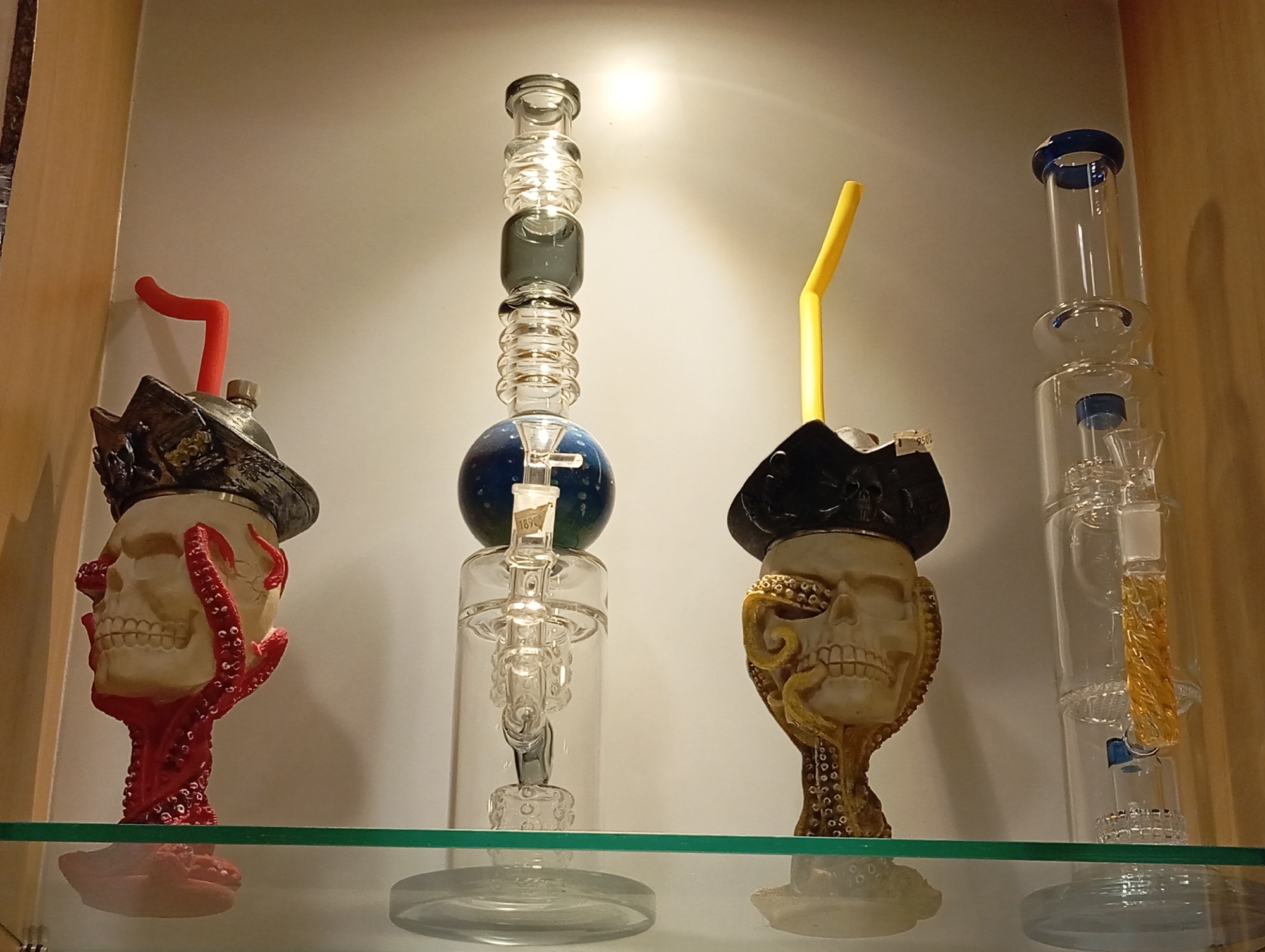 The image shows a shelf in a shop in Karon, Thailand, displaying four glass smoking devices. Two of the devices have skull-shaped bases adorned with tentacle-like embellishments, one red and one gold. A third device has a spherical, blue-colored section and a yellow straw-like attachment. The fourth device is a tall, clear glass cylinder with blue accents. The items are illuminated by a light source above, casting shadows on the wall behind them. The shelf is made of clear glass.