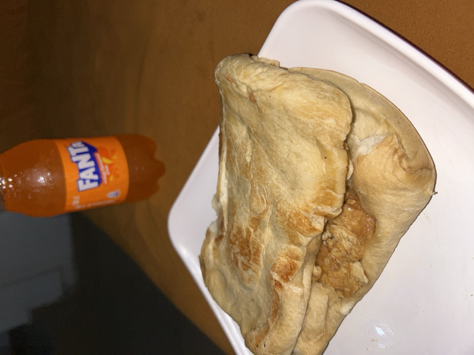 In the city of Egbe, Nigeria, a close-up, elevated view captures a meal setup on a brownish-orange surface. The primary subject is a food item, resembling a flatbread sandwich or wrap, positioned on a white, rectangular plate with rounded edges in the foreground. The flatbread exterior is light beige, with irregular brown areas from cooking. Visible through an opening in the bread is a filling consisting of irregular, light brown pieces of cooked protein, consistent with chicken or similar meat. In the background, slightly out of focus and to the right, stands a small, transparent plastic bottle of Fanta orange-flavored soda. The bottle displays condensation on its exterior and features the distinct orange and blue "FANTA" logo. The ambient lighting suggests an indoor setting. No individuals or active interactions are present in the frame.