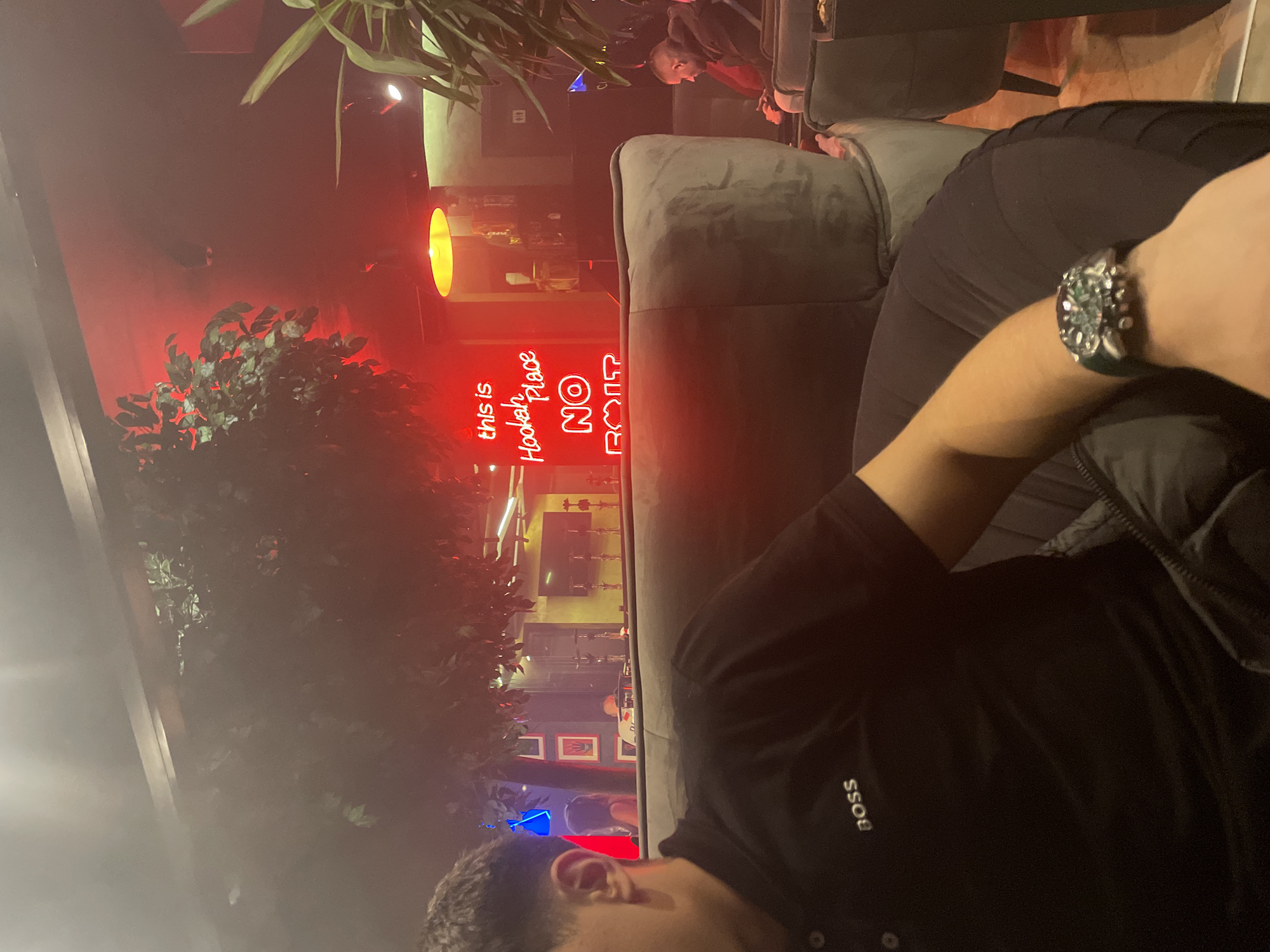 The photo appears to be taken inside a "Hookah Place NO ROAR" establishment, likely in Varna, Bulgaria, based on the presence of Cyrillic characters and the establishment's name. A person wearing a BOSS-branded black shirt and a watch is sitting on a gray velvet sofa. Behind them, a red neon sign reads "this is Hookah Place NO ROAR". Further in the background, a yellow light fixture is visible. Other people are present in the space, sitting in chairs. Plants are visible, one in the foreground partially obscuring the view of the interior, and another seemingly potted near the sign. The environment appears to be dimly lit, with red lighting prominent.