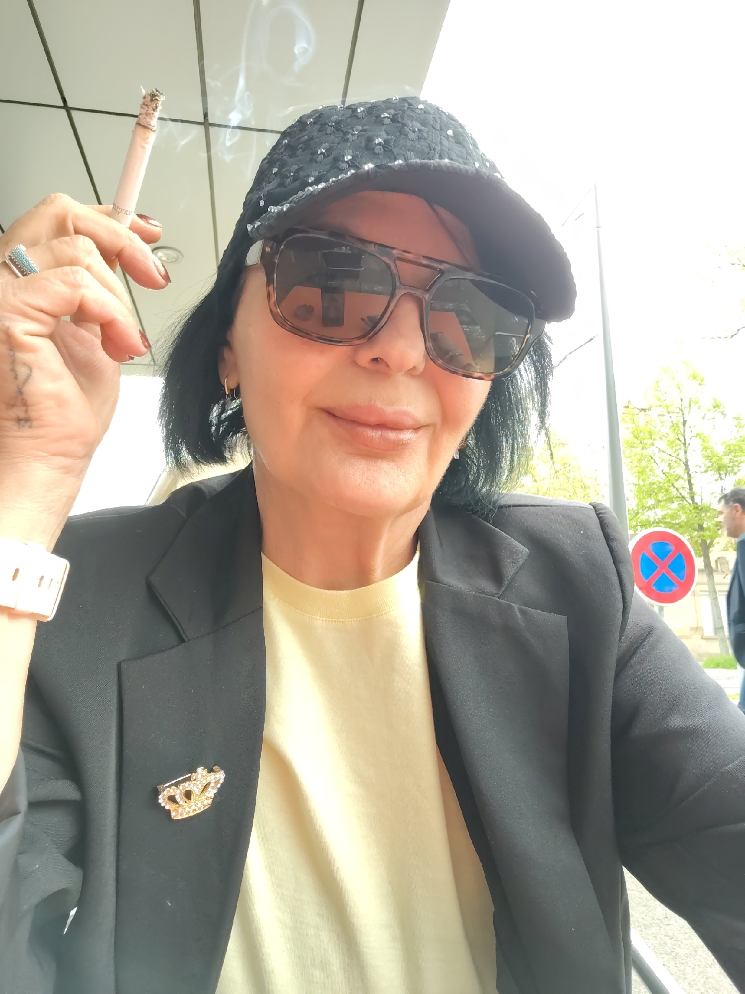 A woman is sitting outdoors, holding a lit cigarette in her left hand. She is wearing sunglasses, a dark-colored cap with decorations, a yellow shirt, and a black blazer with a gold crown-shaped pin. She has dark hair and a tattoo on her left hand. A blue and red parking sign is visible in the background, and a man is walking in the distance. The location is likely in Strasbourg, France, based on the style of the signage and architecture.