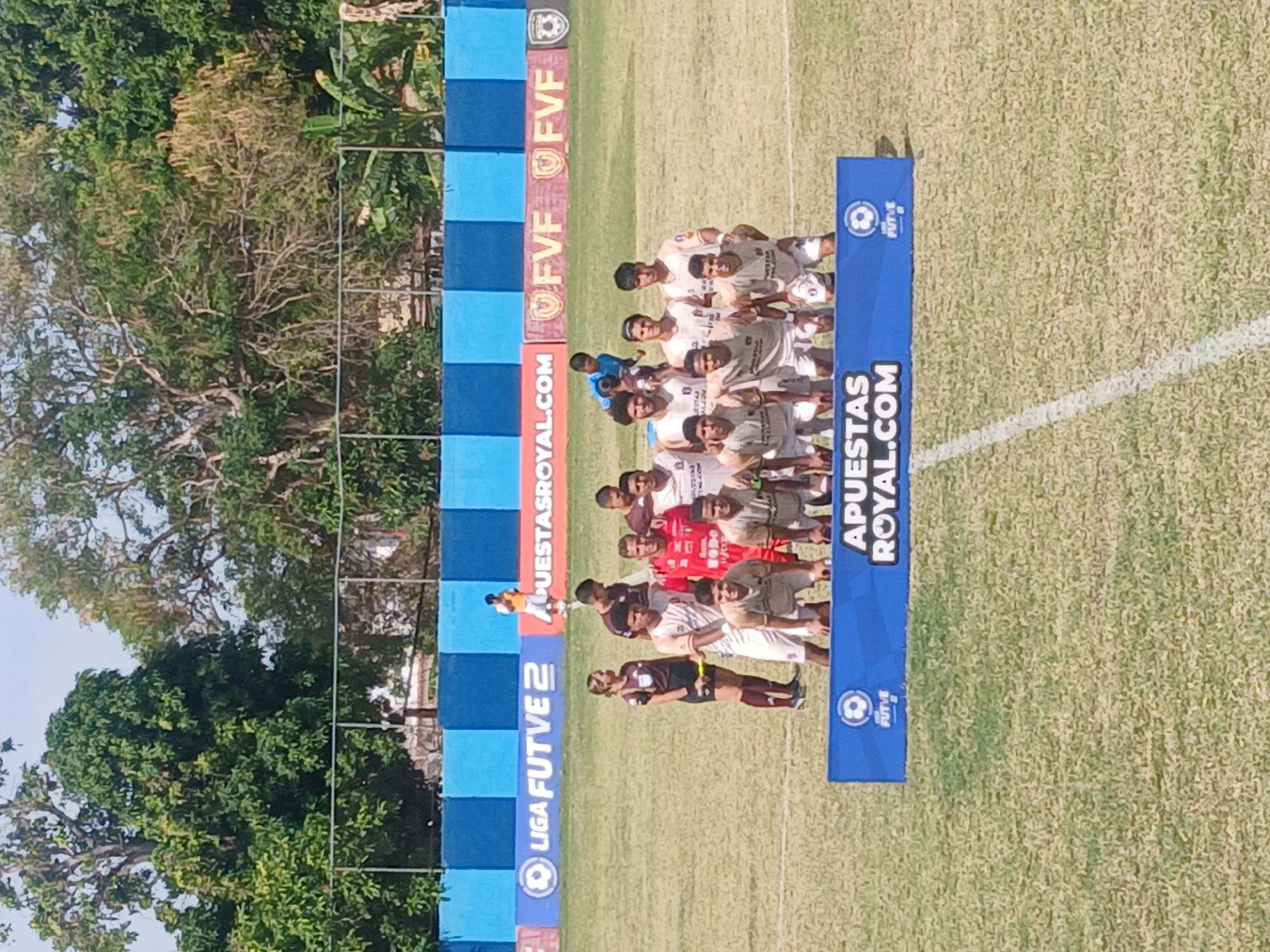 The image depicts a soccer team posing for a team photo on a grassy field in Ciudad Bolivar, Venezuela. There is a diverse group of approximately 15 players and referees dressed in varying uniforms. A set of banners surrounds them, with sponsors identified by logos such as "LIGA FUTVE 2", "APUESTAS ROYAL.COM", and "FVF". The surrounding environment includes trees and a clear sky, indicating a daytime setting.