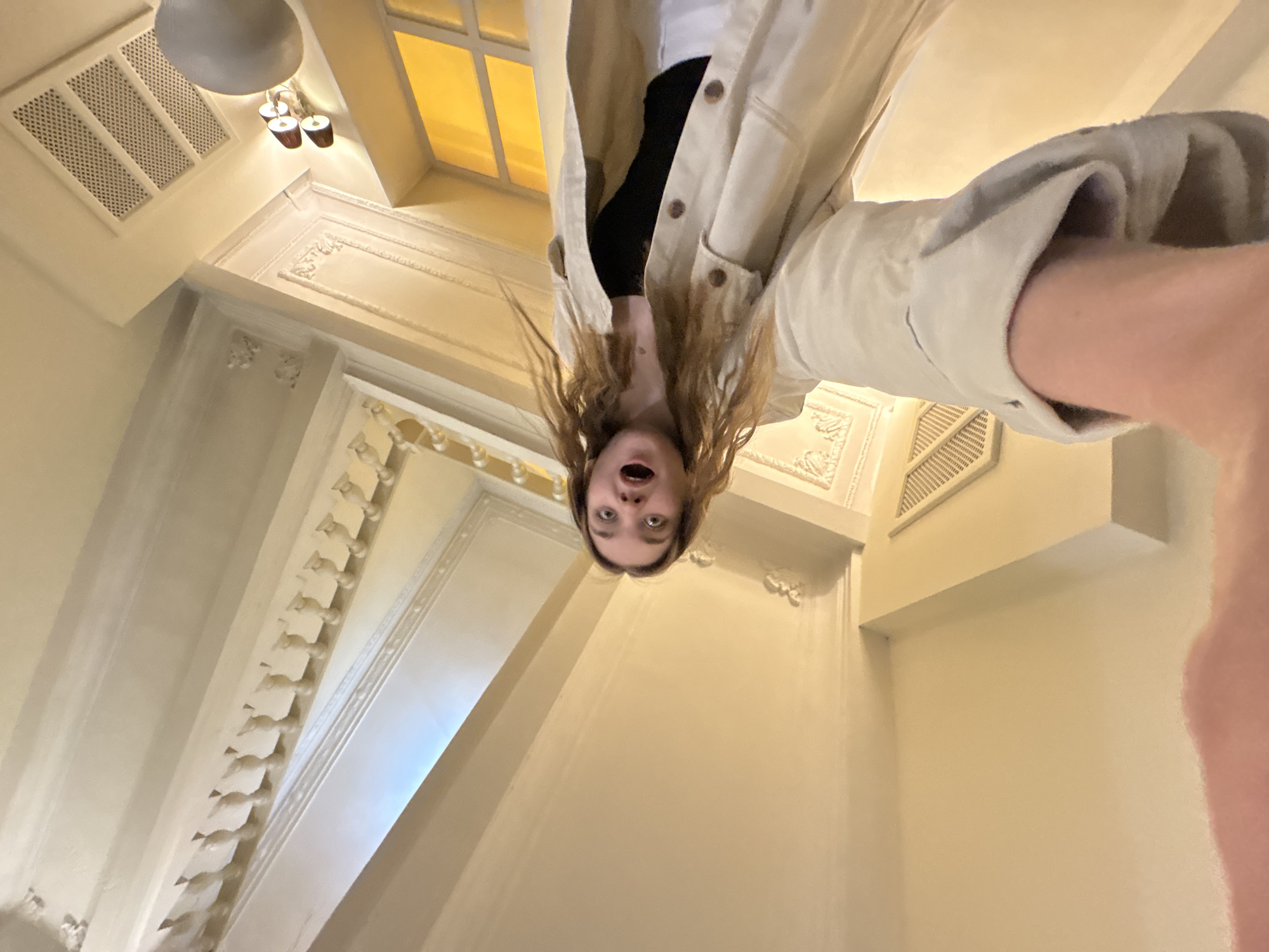 The image shows a young woman with brown hair wearing a light-colored jacket and a black top, with her mouth open in surprise or a yell. She is taking a selfie from a very low angle, pointing the camera upwards. The background appears to be the interior of a building, likely a hallway or stairwell with architectural details. The walls are light-colored and decorated with ornamental molding, and there are ceiling fixtures visible, including a round lamp and a window. The location is Perm, Russia.