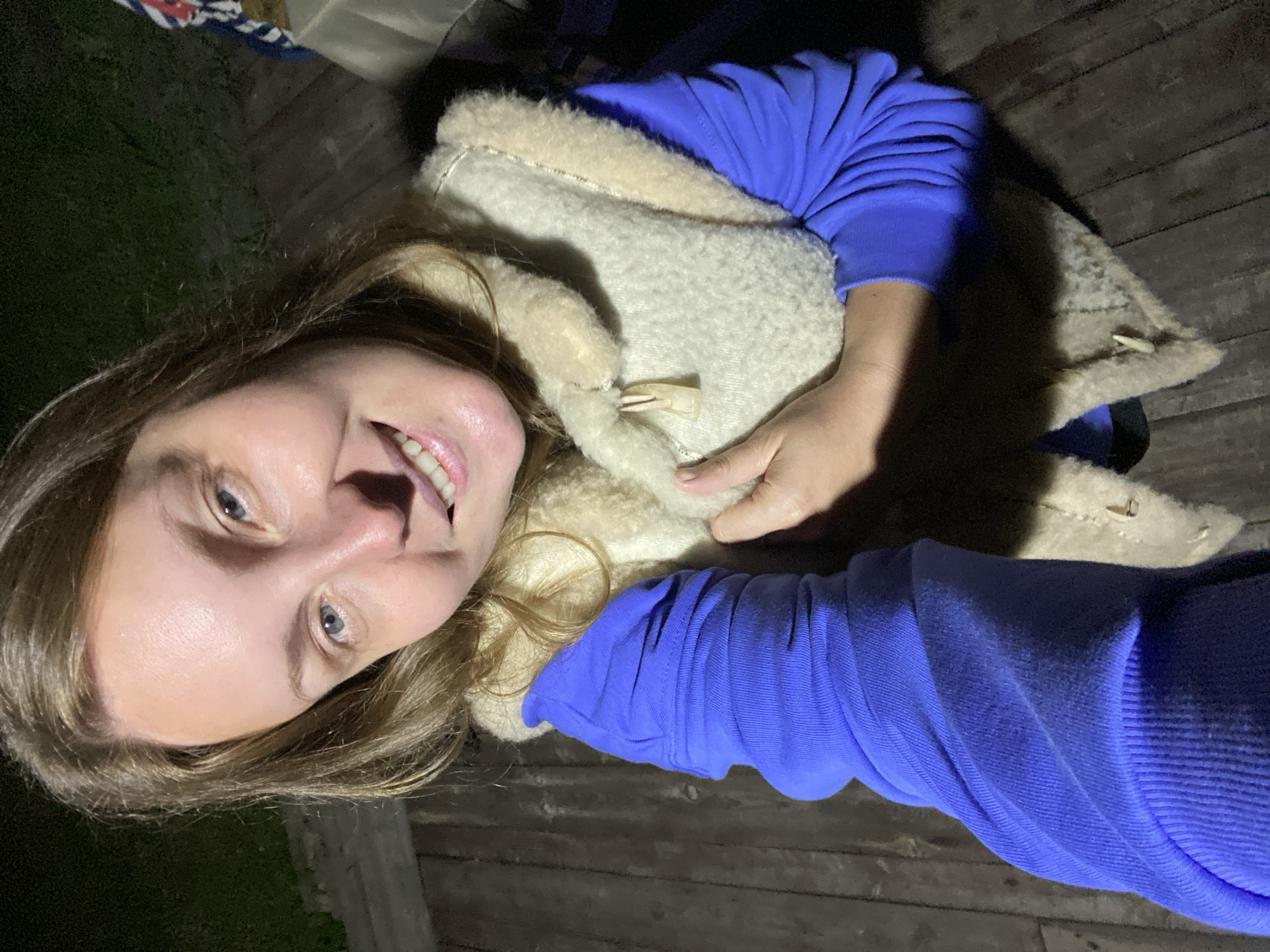 The image shows a woman with light brown hair and blue eyes, smiling at the camera. She is wearing a blue long-sleeved shirt and a cream-colored vest with a fur trim and a wooden toggle closure. The woman is holding her left arm with her right hand, which is partially visible. She appears to be outdoors on a wooden deck or porch at night, as indicated by the dark background and the lighting on her face. There is some greenery visible in the background, as well as a table with items on it. The location is specified as Garevaya, Russia.