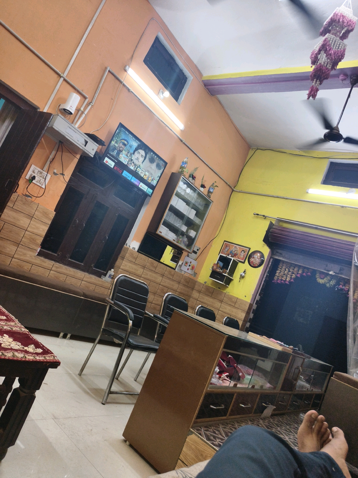 The image captures an interior view of what appears to be a shop or small office in Budhana, India. The walls are painted in contrasting orange and yellow. In the foreground, a person's bare feet are visible, indicating they are likely seated or lying down. Several black office chairs with metal frames are arranged around a long, wooden counter or desk. This counter has a glass display top, beneath which various items are stored in drawers and compartments. The floor is tiled.
On the orange wall, a television screen displays a movie poster with actors. Above the TV, a white device is mounted. Electrical conduits and switches are visible on the walls.
To the right, a large shop shutter is partially open, revealing a dark interior. Decorative garlands hang above the shutter. On the yellow wall, framed religious pictures are displayed, suggesting a cultural or spiritual element to the space. A ceiling fan is visible, and a decorative hanging ornament is suspended from the ceiling. The overall environment suggests a functional, possibly commercial space with some personal touches.