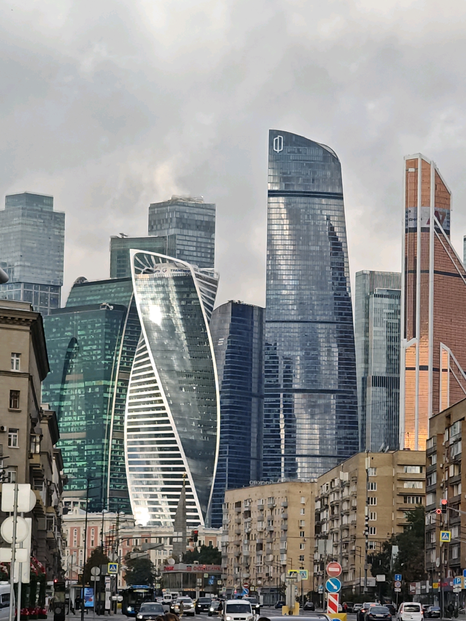 The image shows a street scene in Moscow, Russia, with a prominent view of the Moscow International Business Center (Moscow City). In the foreground, a street lined with apartment buildings and a few businesses is visible. Cars and traffic signs are present on the street. In the background, several modern skyscrapers, characteristic of Moscow City, dominate the skyline. These skyscrapers vary in height and architectural style, with glass facades reflecting the overcast sky. The overall impression is a blend of older urban architecture and contemporary high-rise development in a bustling city environment.