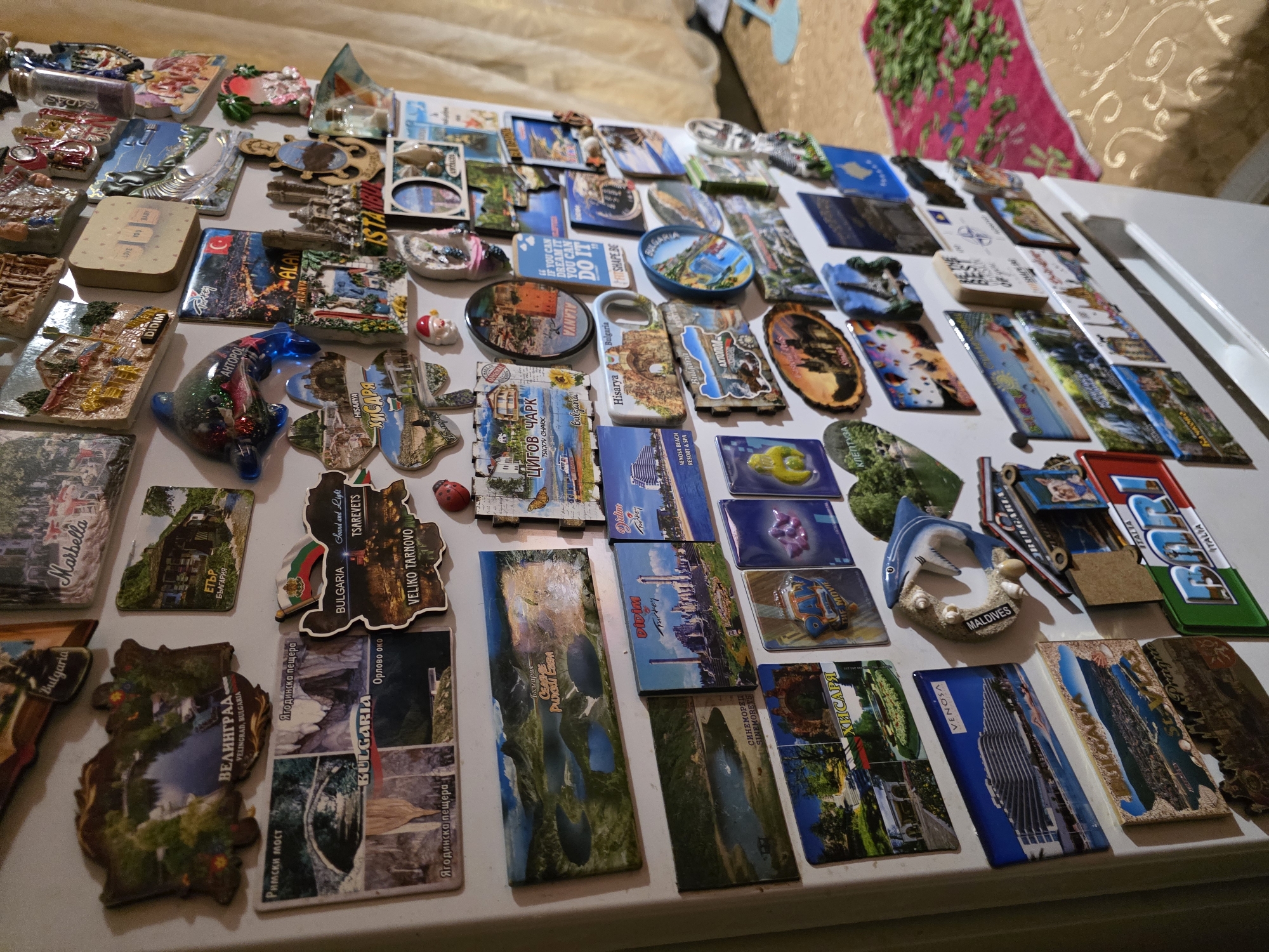 The image shows a collection of souvenir magnets spread out on a white surface. The magnets depict various locations and landmarks from different places, with a notable concentration of magnets from Bulgaria, including places like Veliko Tarnovo, Tsigov Chark, Velingrad, Hisarya, and destinations along the Black Sea coast. Other magnets represent cities such as Venice, Marbella, and Didim in Turkey. The magnets vary in shape, size, and material, some displaying landscapes, landmarks, or city names. Some of the magnets are of the city of Yagodovo.