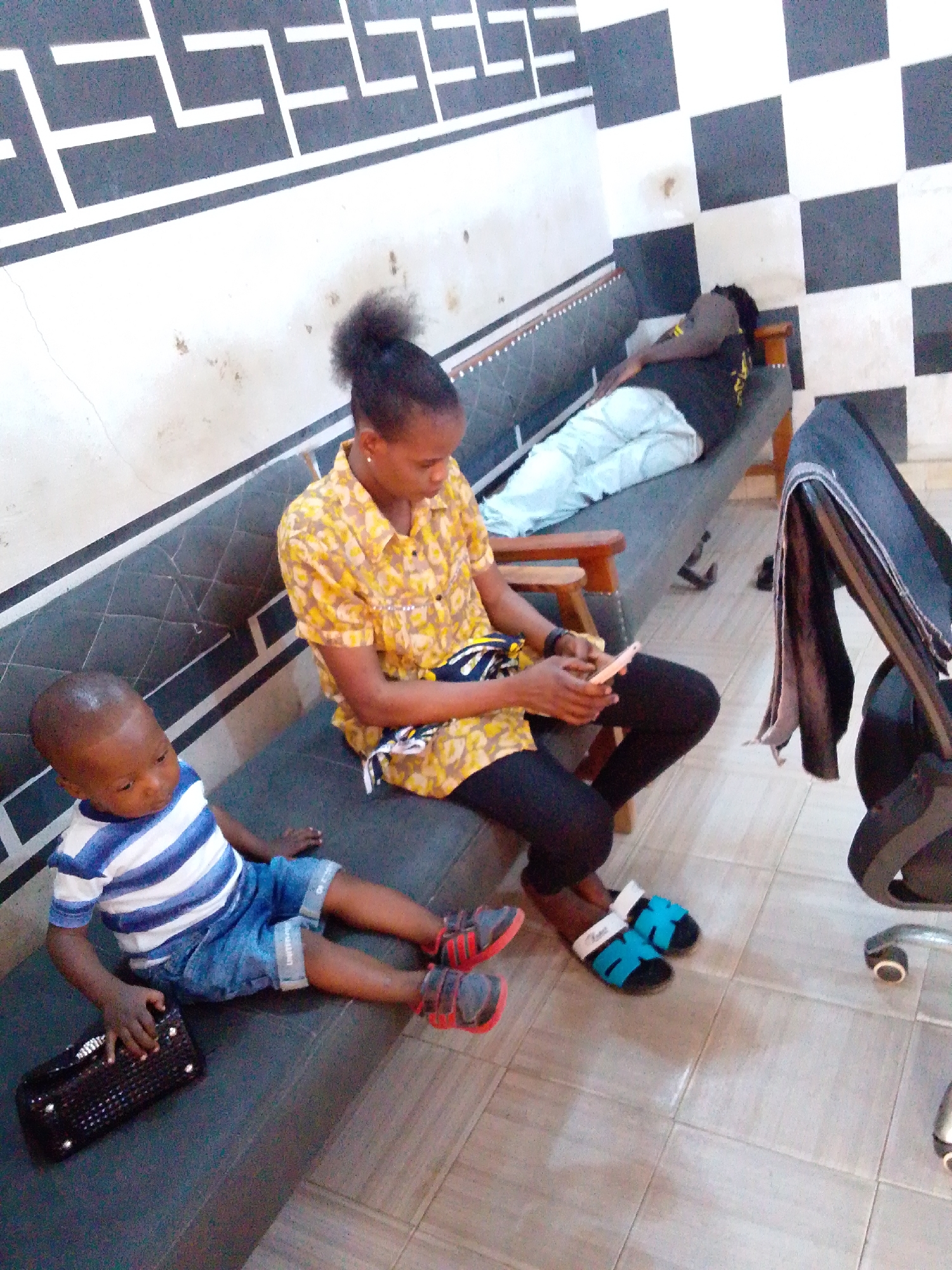 The image depicts an indoor scene. A young child in a blue and white striped shirt and denim shorts is seated on a dark gray cushioned bench holding a dark handbag. A woman with her hair in a bun, wearing a patterned yellow shirt and black pants, is sitting on the same bench looking at a cell phone. Behind them, on a longer bench, a person is lying down. The backdrop is a wall with a geometric black and white design above and checkered black and white tiles to the right. A black office chair is visible on the right side of the image. The location is Unknown, Unknown.
