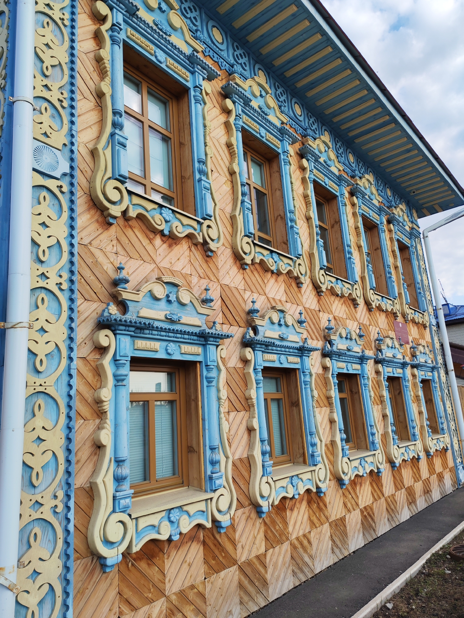 A low-angle, slightly canted, outdoor shot reveals the side of a building in Sarapul, Russia, known for its intricate wooden lace architecture. The exterior of the building is characterized by diagonally placed, light brown wooden planks arranged in a checkered pattern. A row of eight windows stretches across the facade, each framed by elaborate blue and pale yellow wood carvings. The ornate carvings feature curved shapes, floral motifs, and geometric patterns. A light blue drainpipe runs vertically along the left side of the building, and an adjacent carved panel mirrors the window frame detailing. Above the windows, the edge of the roofline is visible, with blue and pale yellow striped accents. The sky is overcast, and a narrow asphalt sidewalk runs along the base of the building. The overall composition emphasizes the detailed craftsmanship of the wooden architecture.