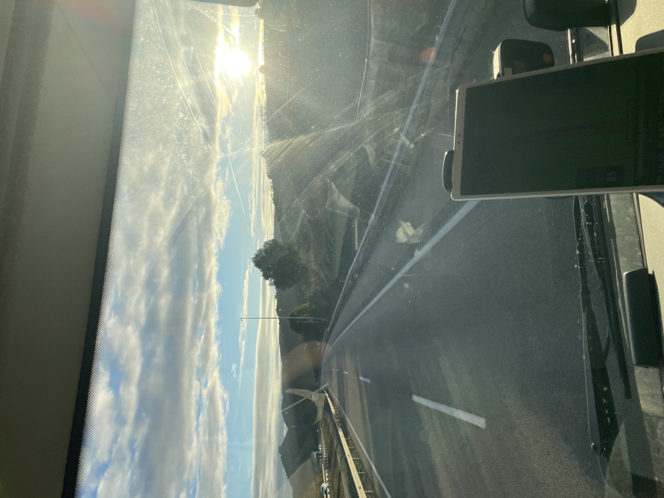 The image presents a view from the interior of a vehicle, looking outward through a dirty windshield onto a highway in Torrente de Cinca, Spain. The foreground features a dark dashboard, the base of windshield wipers, and a white electronic tablet or large smartphone mounted in the lower right, displaying a dark screen with a faint red line. The asphalt highway, marked with white lane lines, extends into the distance. A metal guardrail runs along the right side of the road. The surrounding environment consists of rolling hills covered with green and brown vegetation. A tall light pole stands roadside on the right. A separate road or exit ramp with several vehicles is visible in the far left background. The sky above is partly cloudy with horizontal cloud formations, and the sun is low on the horizon in the upper right quadrant, creating significant glare on the windshield. Faint contrails are visible in the sky. No individuals are visible.