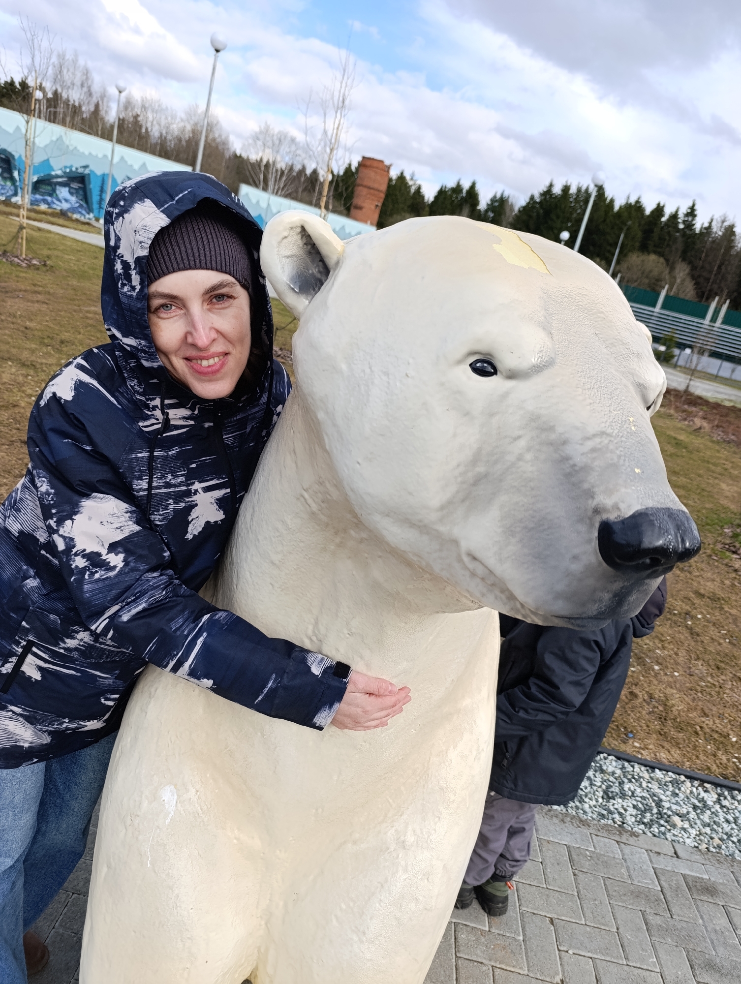 A woman in a blue and white patterned hooded jacket and gray beanie is hugging a large, white polar bear statue. The woman is smiling and looking at the camera. The polar bear statue has a black nose and eyes. The background includes a grassy area, trees, and a brick structure. A child is partially visible behind the polar bear. The location is the city of Perm, Russia.