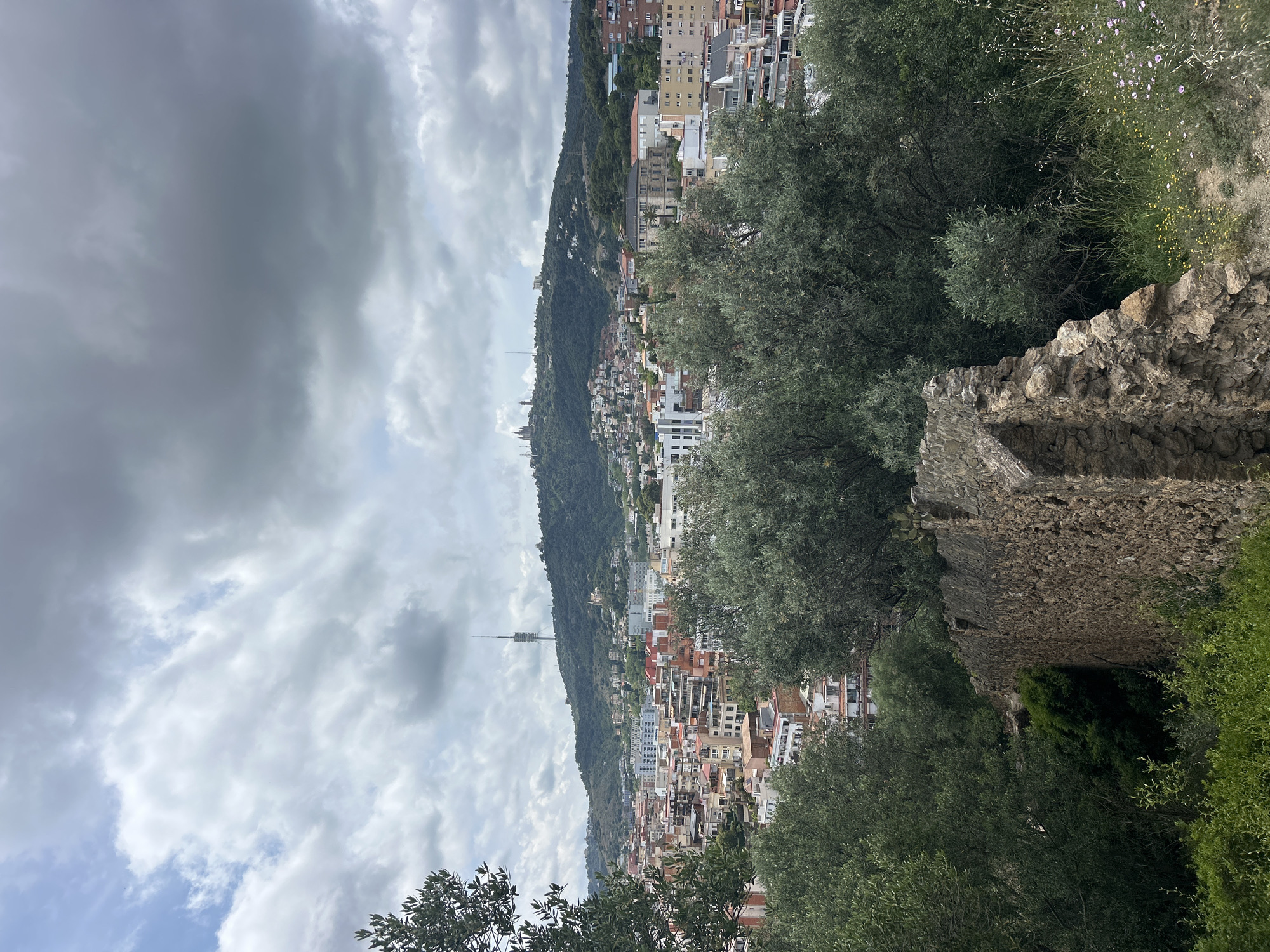 The image depicts a view of Barcelona, Spain, likely taken from a hillside vantage point. The foreground features trees and a stone wall. In the midground, a densely populated cityscape stretches along a hillside, with buildings of various heights and colors. A tall telecommunications tower is visible. The sky is overcast with heavy clouds.